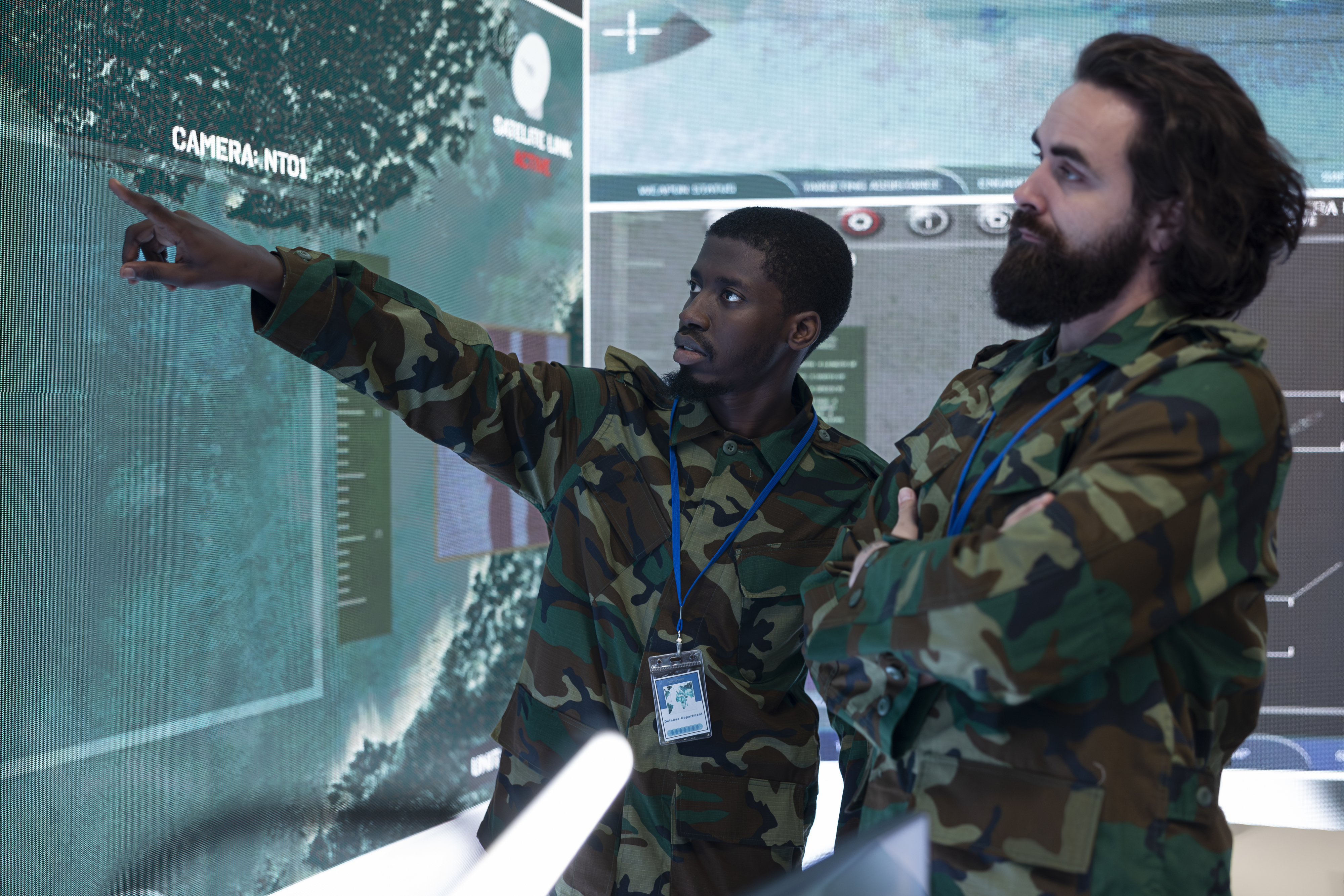 Two individuals in military camouflage uniforms discuss tactical details on a large digital screen