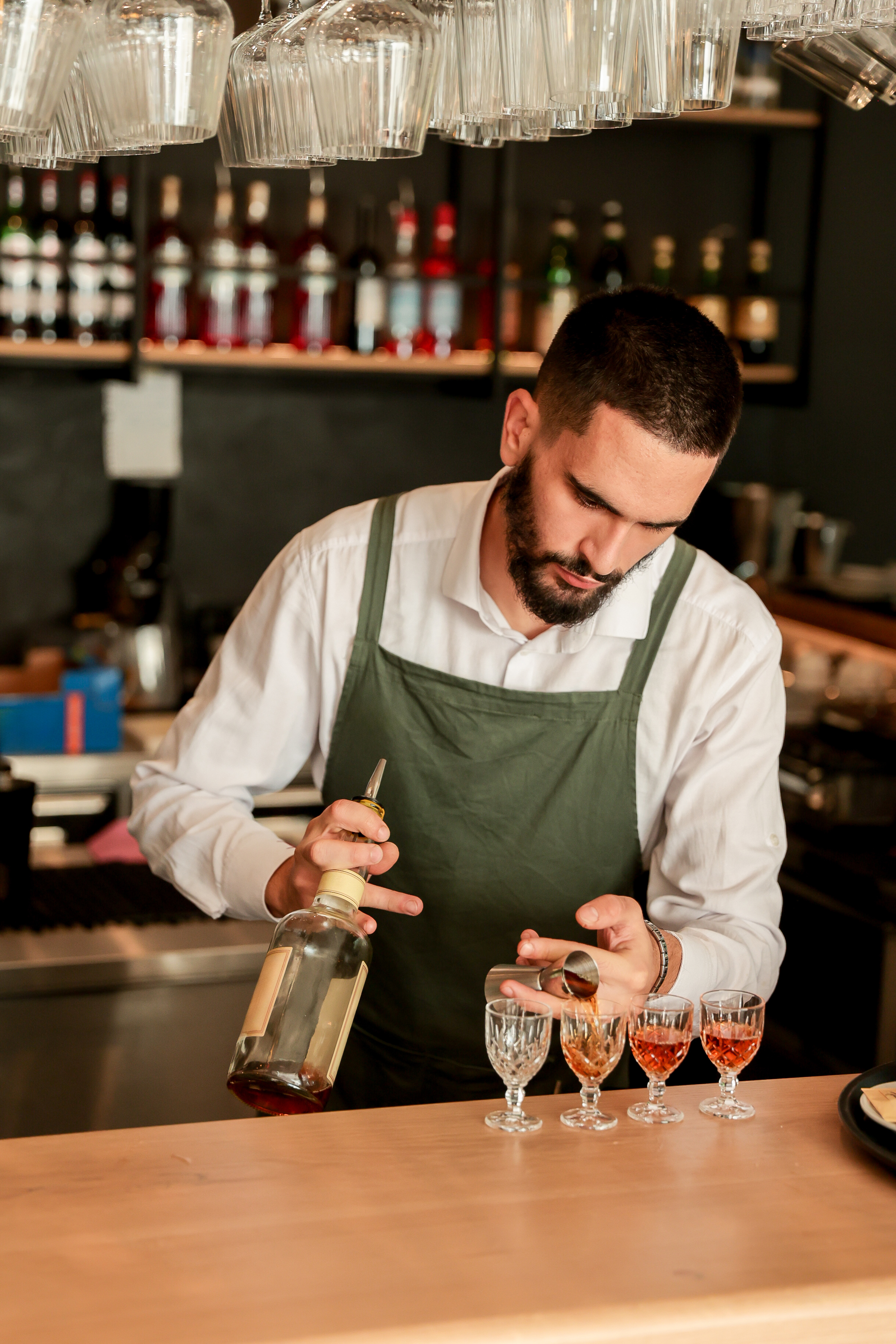 Bartender wearing a green apron is pouring and measuring drinks into four glasses at a bar