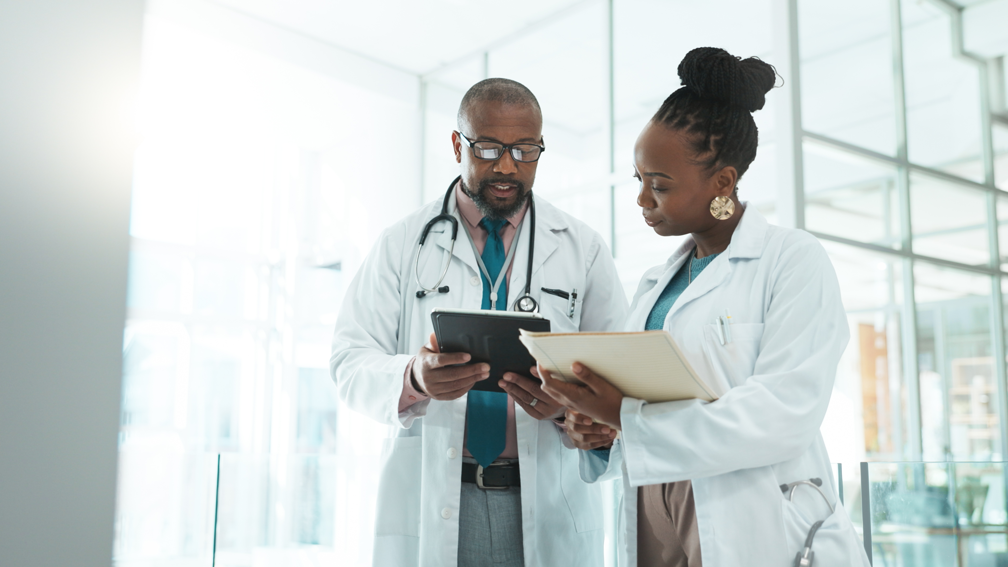 Two doctors, one holding a tablet and the other a folder, discuss in a hospital corridor. They&#x27;re both dressed in white lab coats