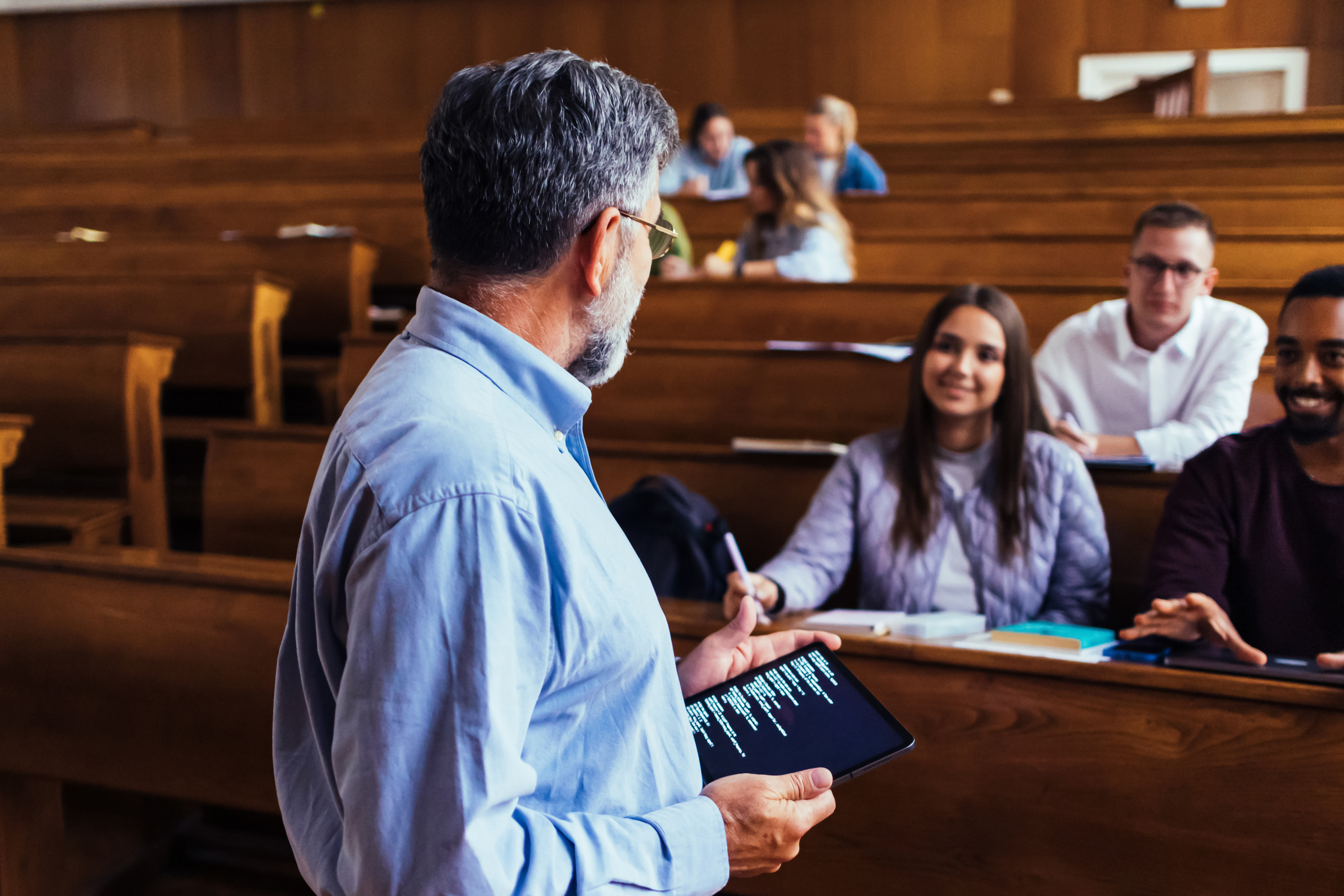 Professor lecturing a diverse group of students in a classroom, holding a tablet while students take notes and listen attentively