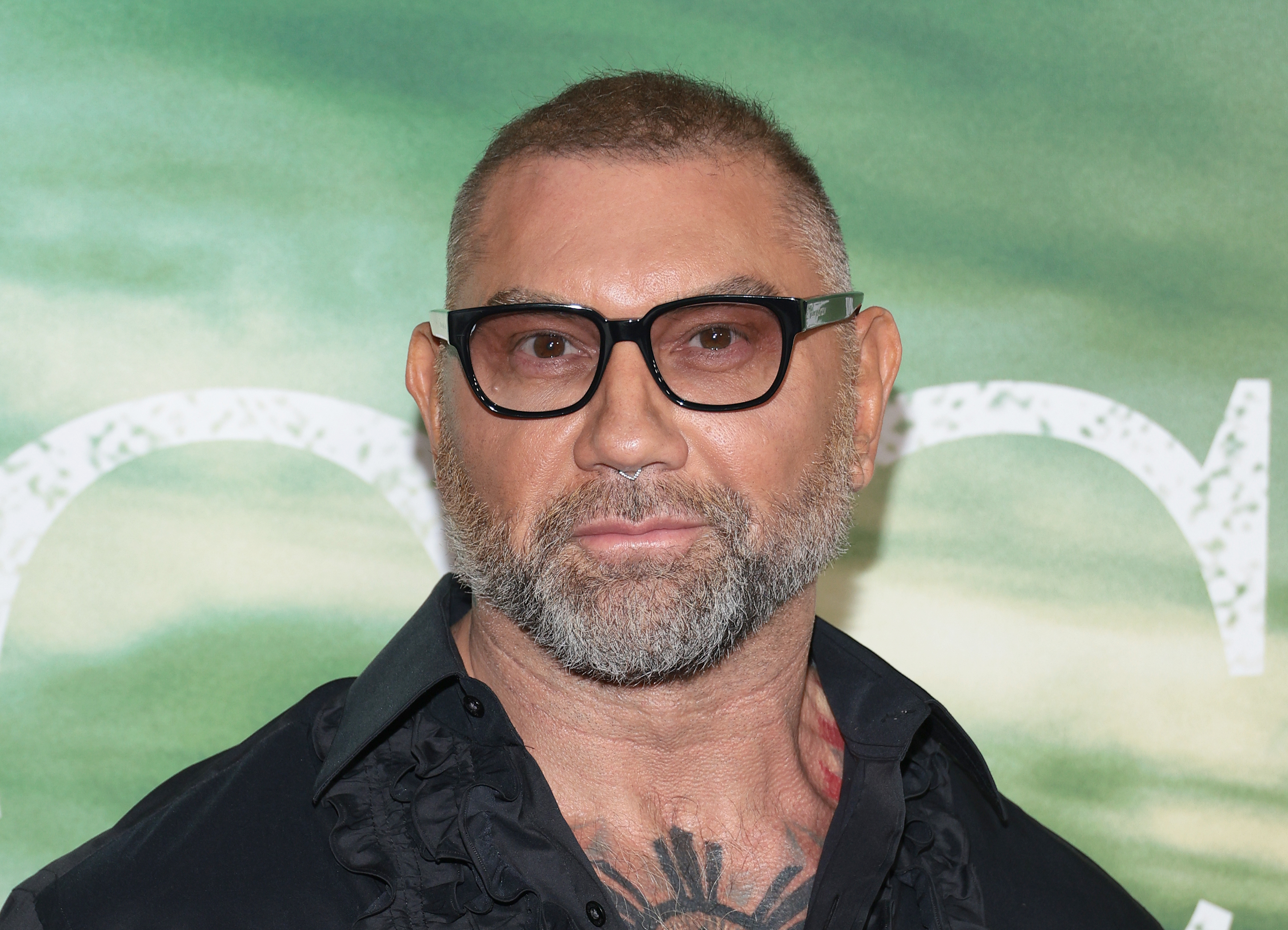 Person in glasses with a short beard, wearing a dark shirt with ruffled details. Green background suggests an event setting