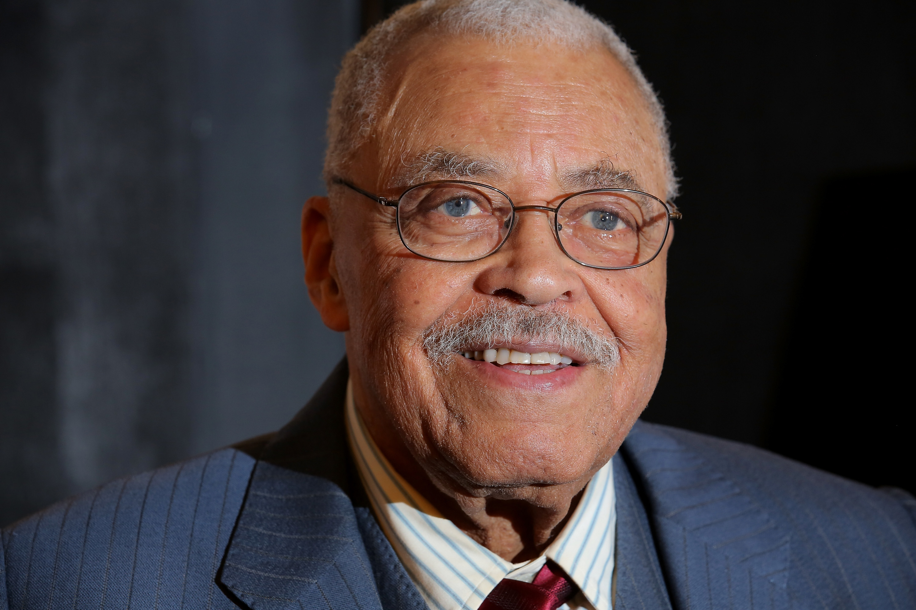 Elderly man in a suit and tie, smiling, with glasses and a mustache, against a dark background