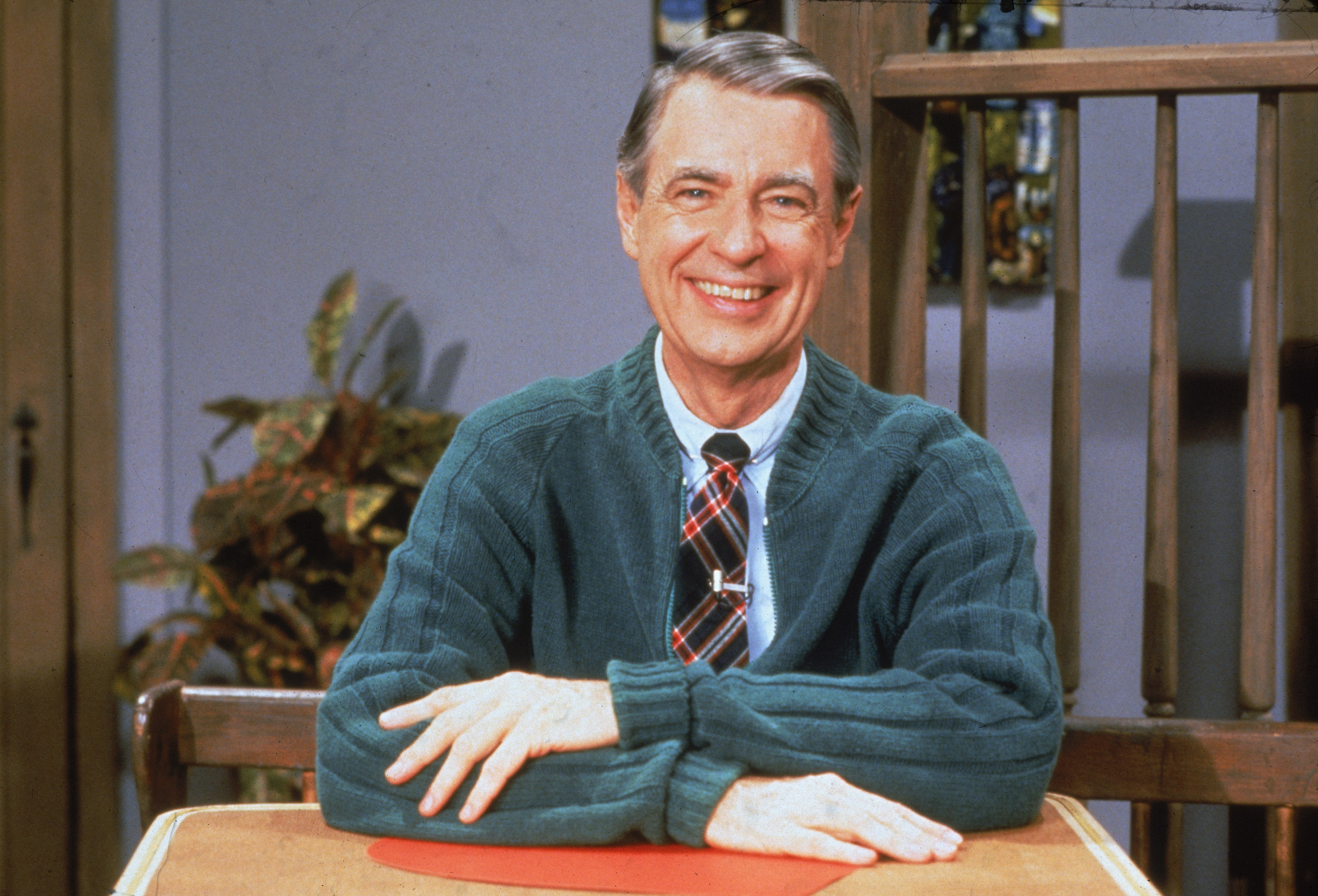 A person smiles warmly while seated at a table, wearing a cardigan and plaid tie, reminiscent of a classic TV show host