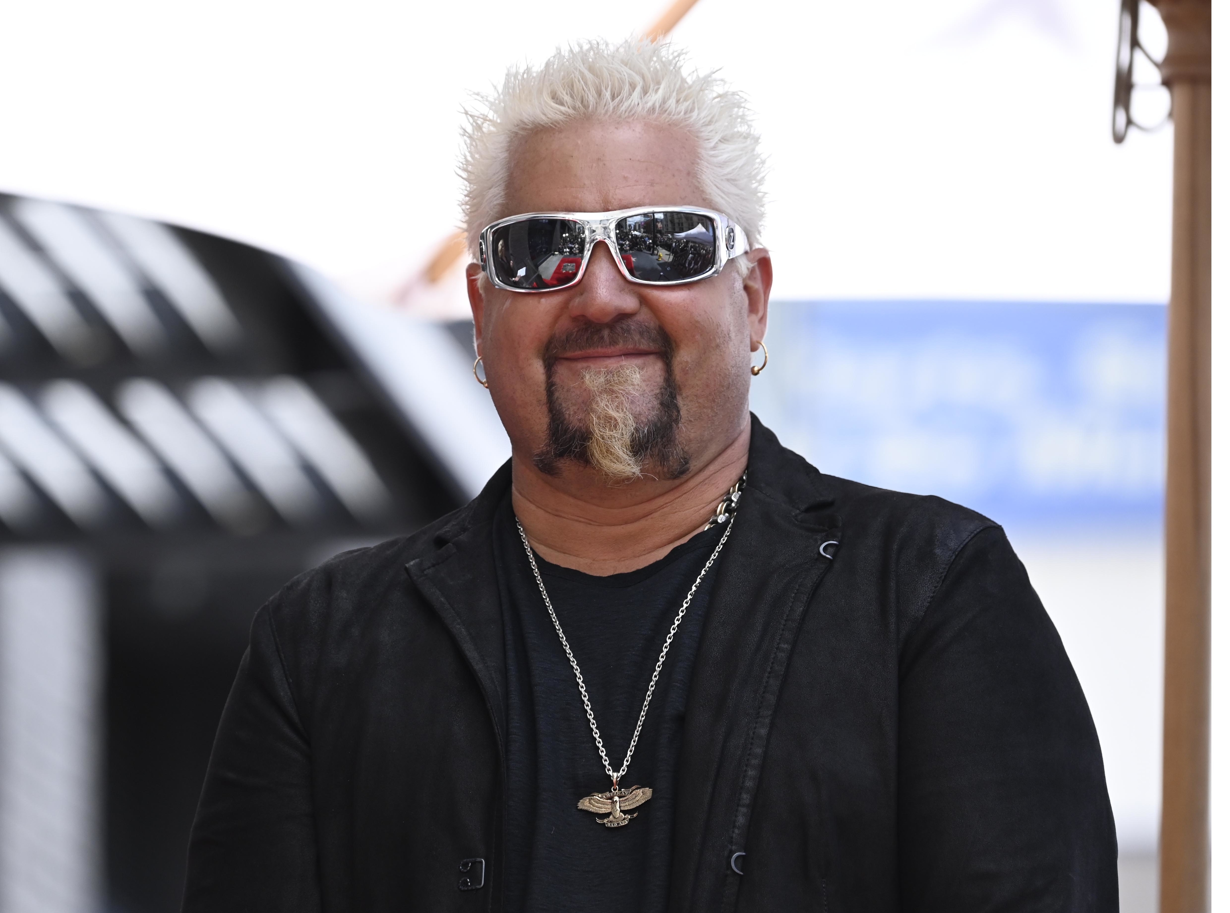 Person with spiky hair and sunglasses wearing a black jacket and chain necklace