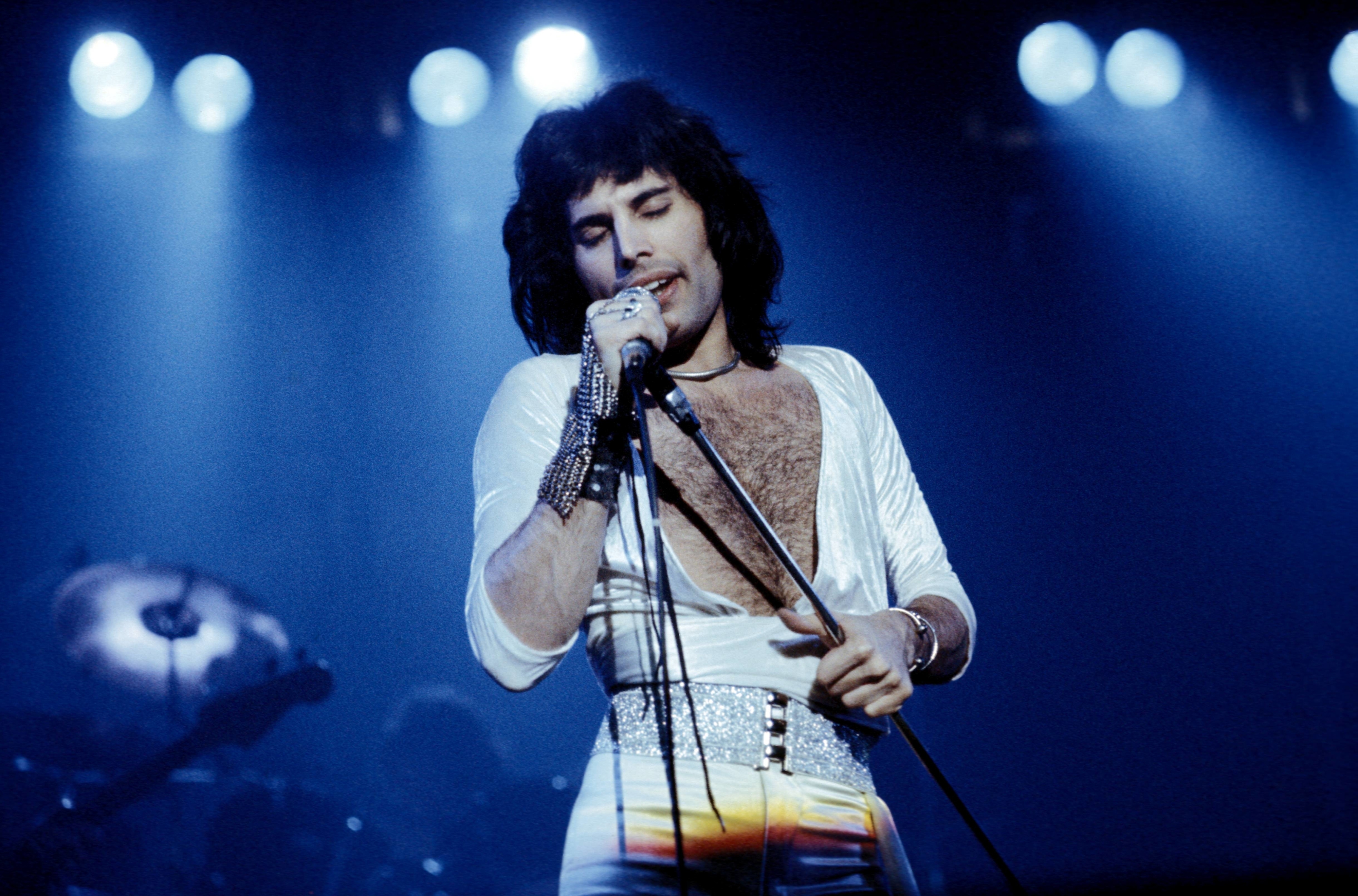 Singer performing passionately on stage, wearing a deep V-neck top and glittery belt, holding a microphone. Stage lights shine in the background