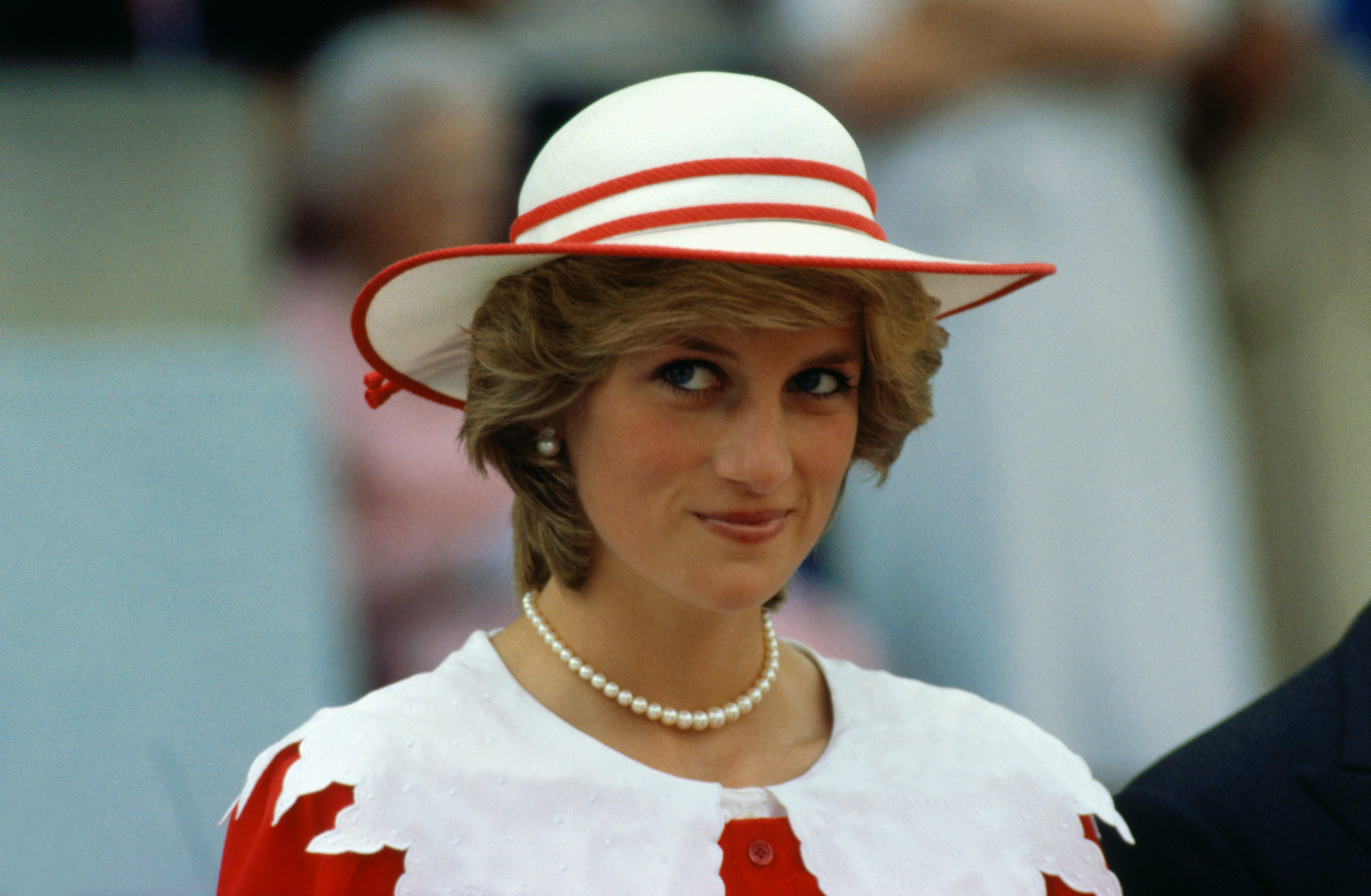 Woman wearing a hat with a red band and a pearl necklace, looking at the camera with a slight smile