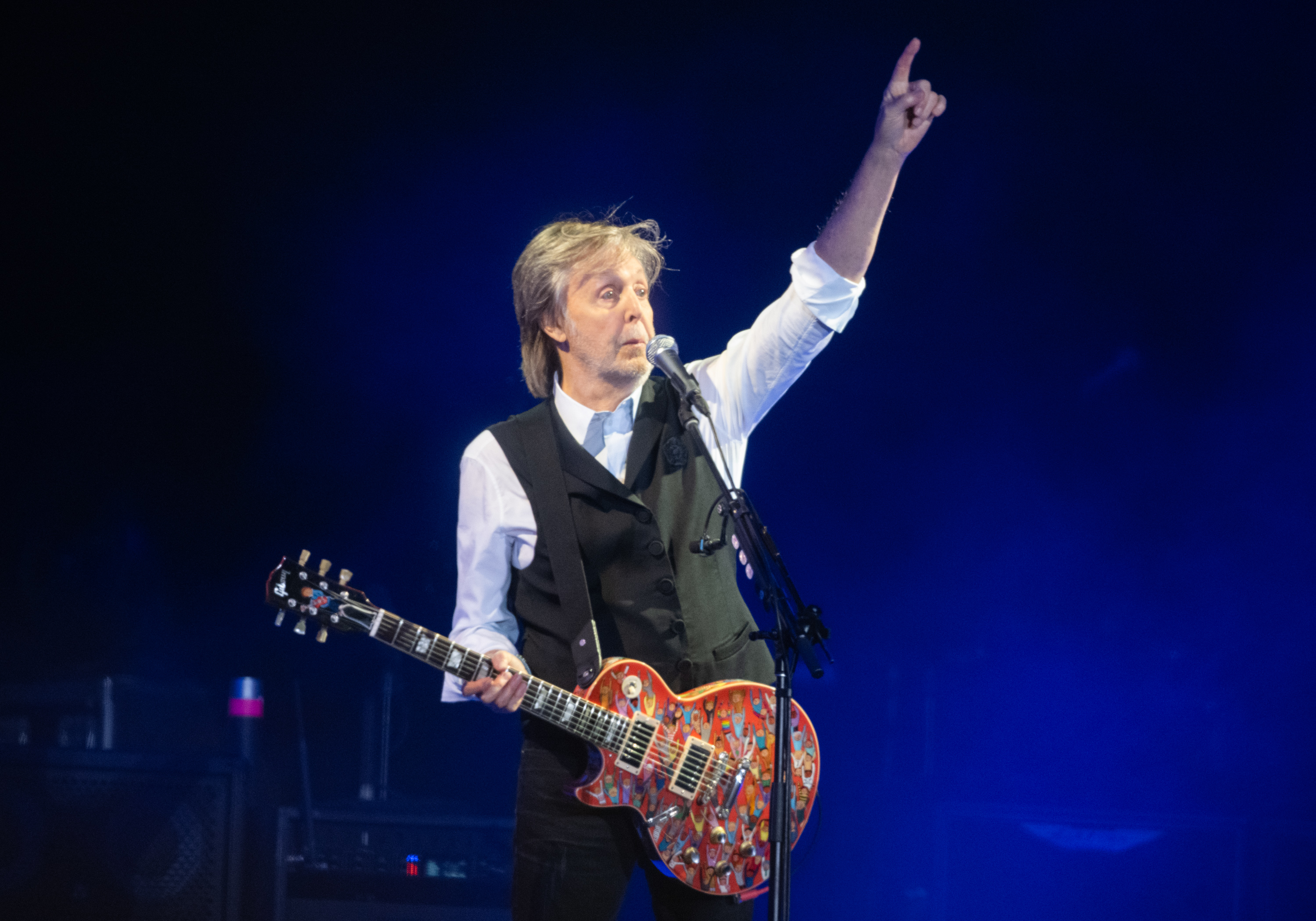 Musician playing guitar on stage, wearing a white shirt and black vest, pointing upwards