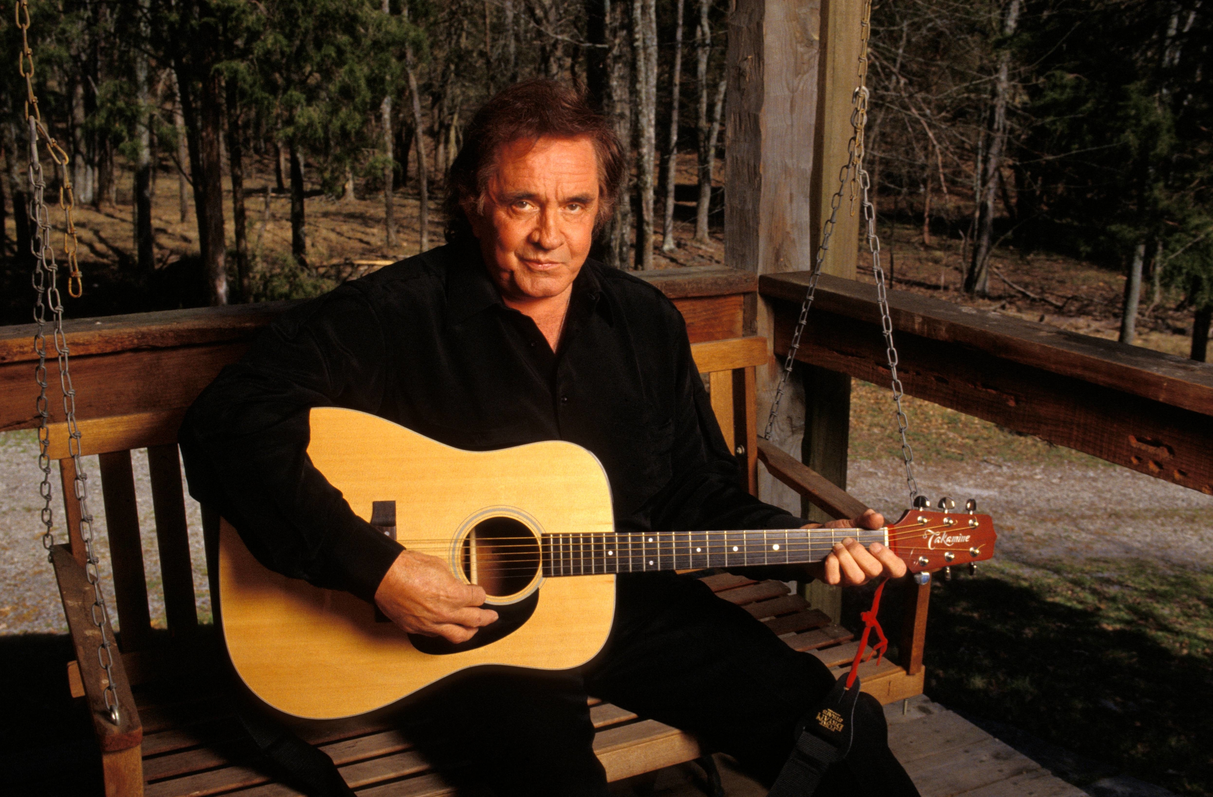 A person sits on a porch holding an acoustic guitar, wearing a dark-colored shirt. The setting is outdoors with trees in the background