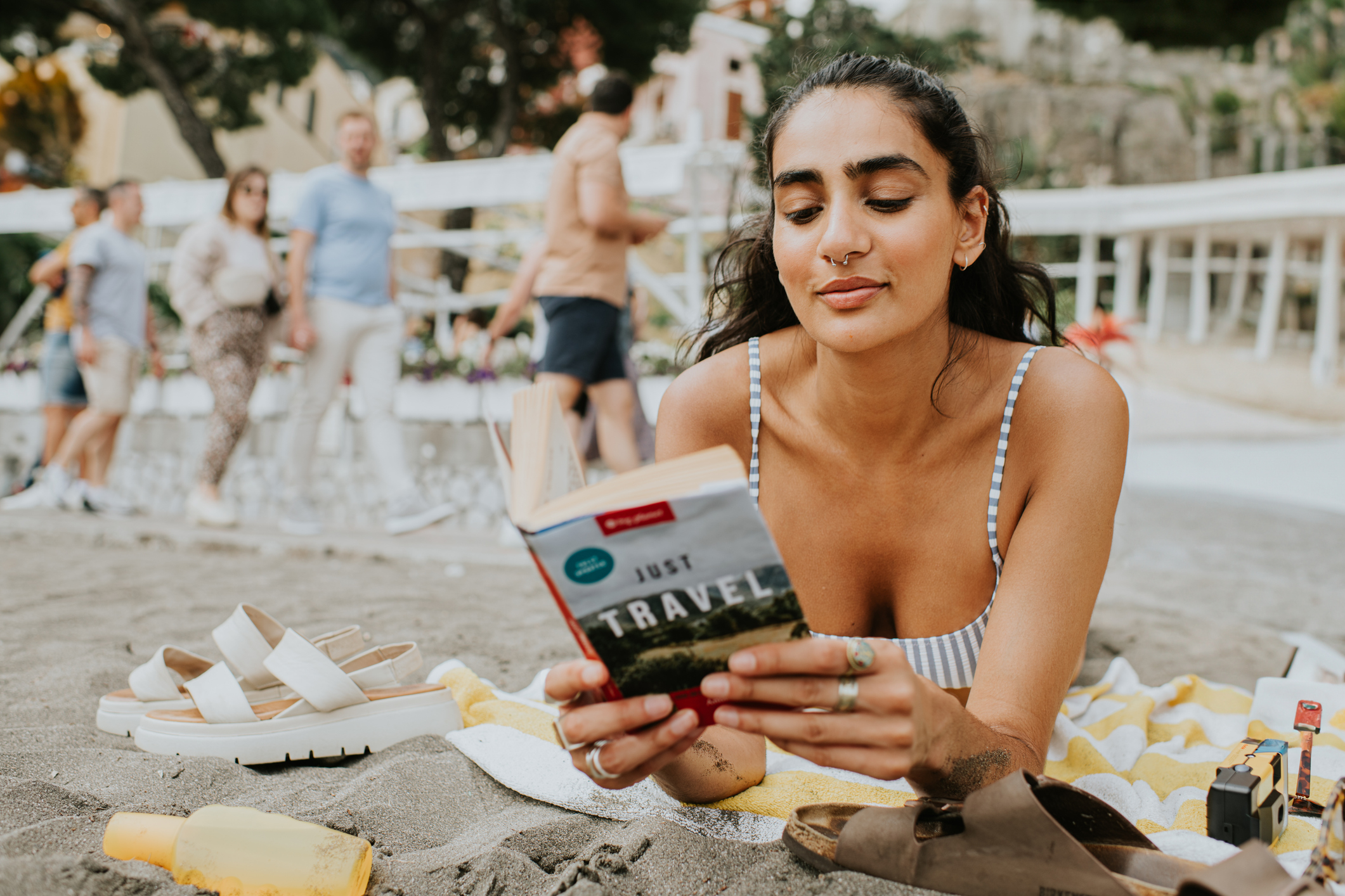 A woman lies on the beach reading a travel book. Sandals and a drink are beside her. People in the background are walking