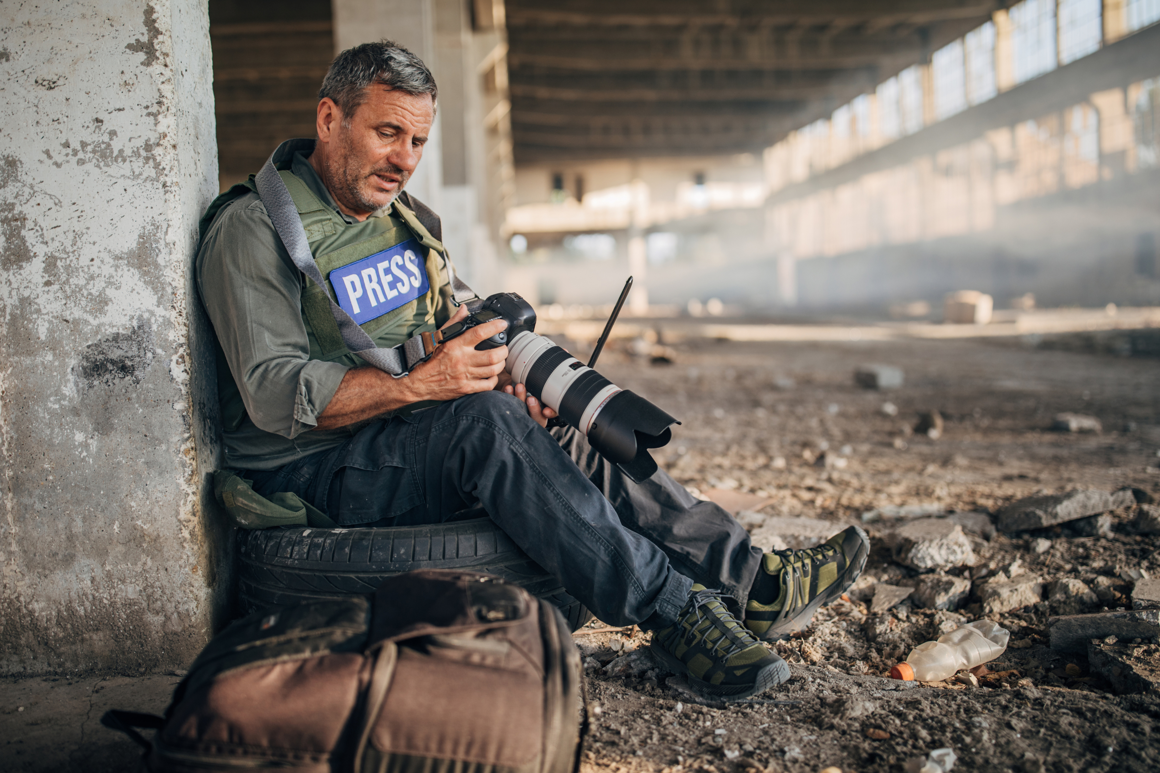 A journalist with a &quot;Press&quot; vest sits in a deserted building, holding a camera with a large lens, surrounded by debris