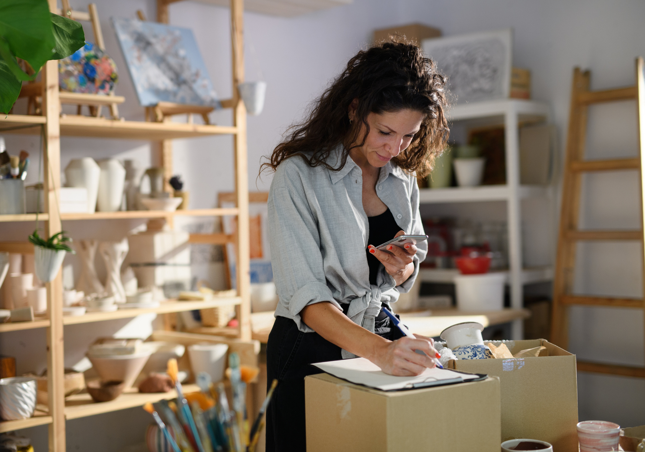 Woman in a pottery studio, holding a phone and writing on a notepad atop a cardboard box. Shelves with pottery and art supplies in the background