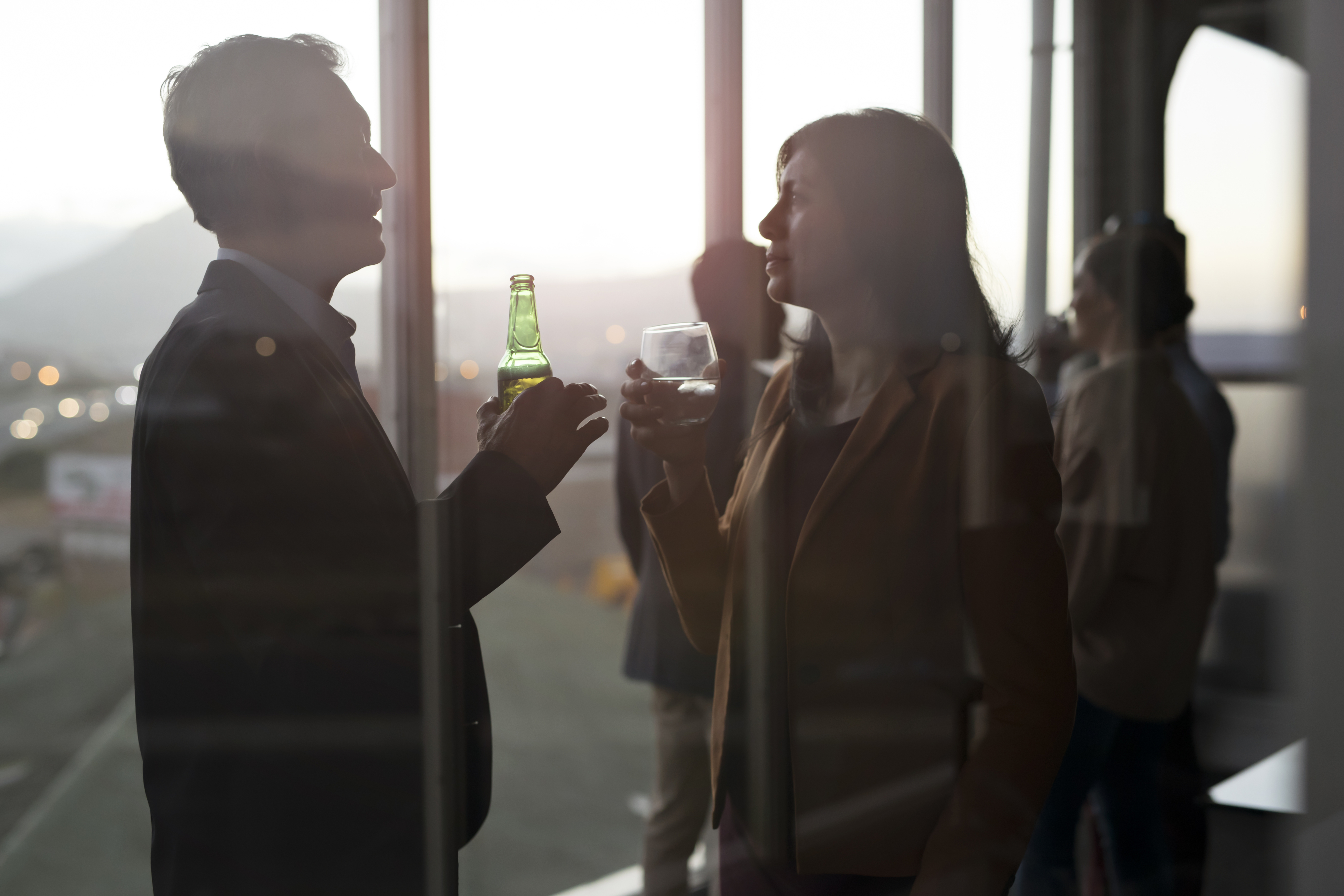 Two people in formal attire are silhouetted against a window, holding drinks, and engaged in conversation at a social event