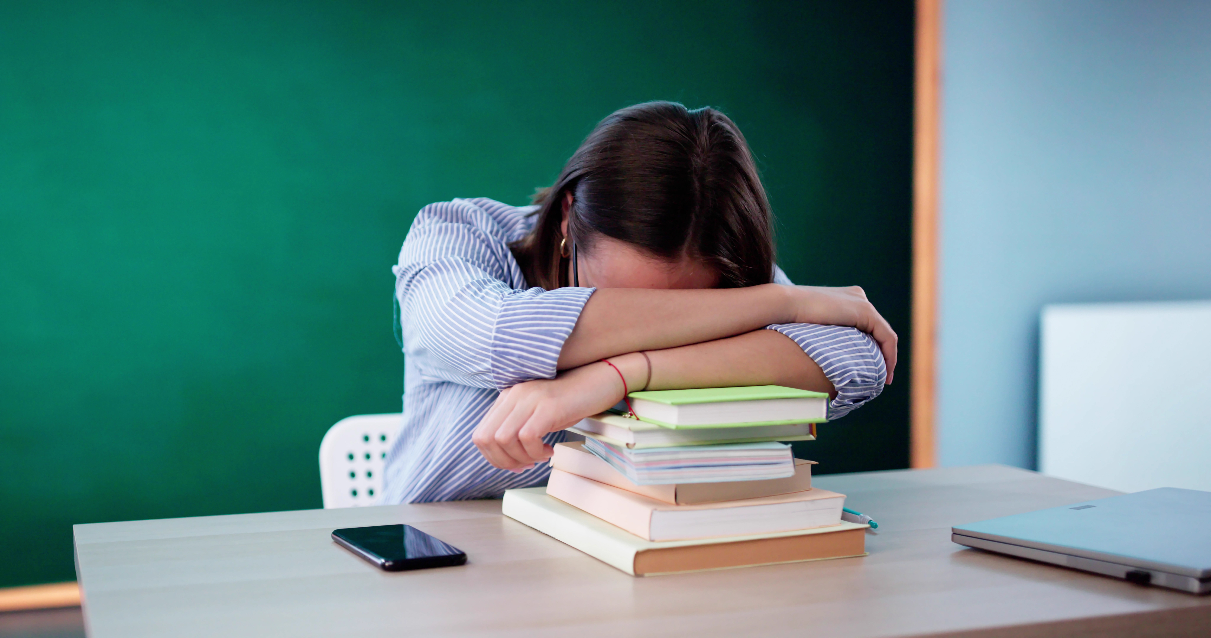 Person with head on arms over stacked books on desk; appears exhausted or overwhelmed