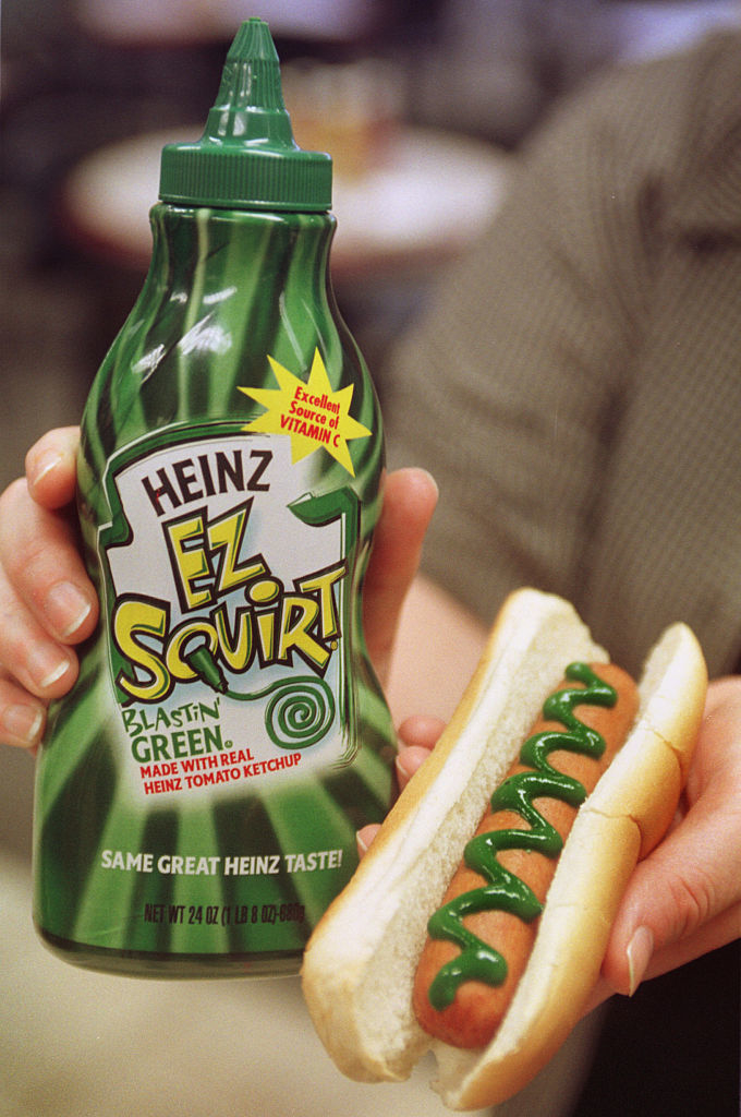 Person holding Heinz EZ Squirt &quot;Blastin&#x27; Green&quot; ketchup bottle and hot dog with green ketchup drizzle