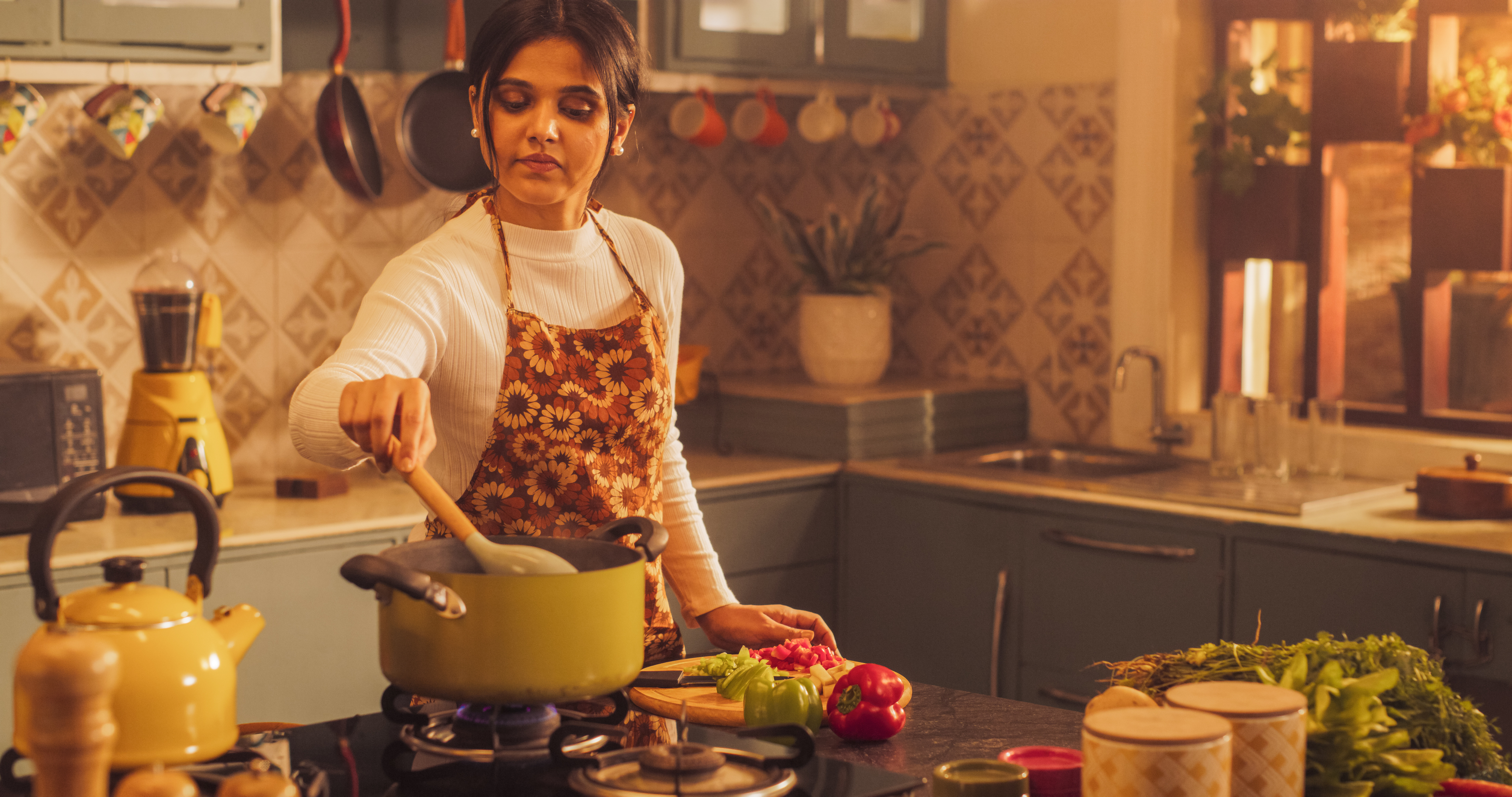 Person cooking in a cozy kitchen, stirring a pot on the stove. Vegetables and kitchenware are visible on the counter