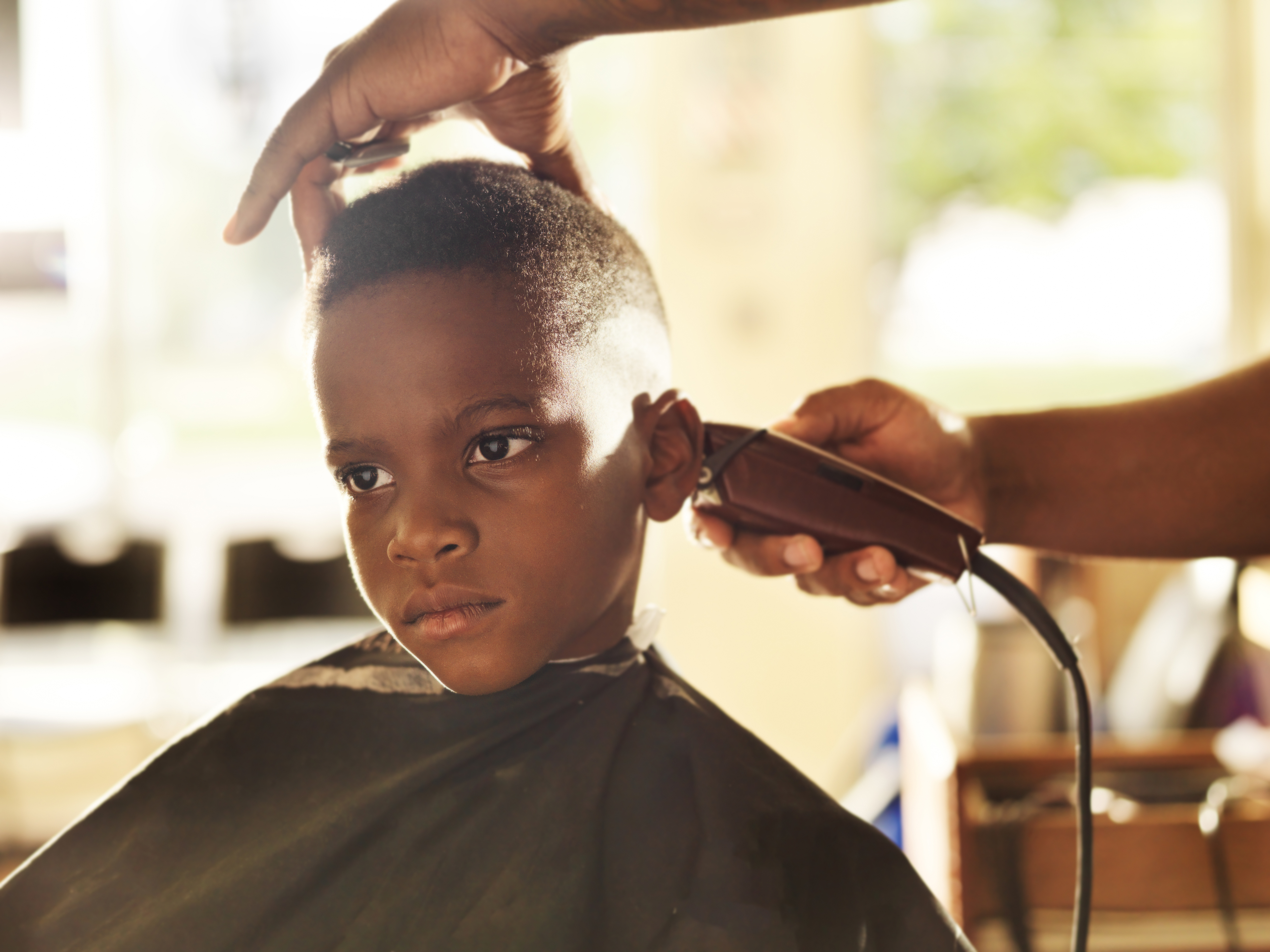 A child receives a haircut at a barbershop, viewed with a focused expression. Hands of the barber are visible, holding clippers and adjusting the hair