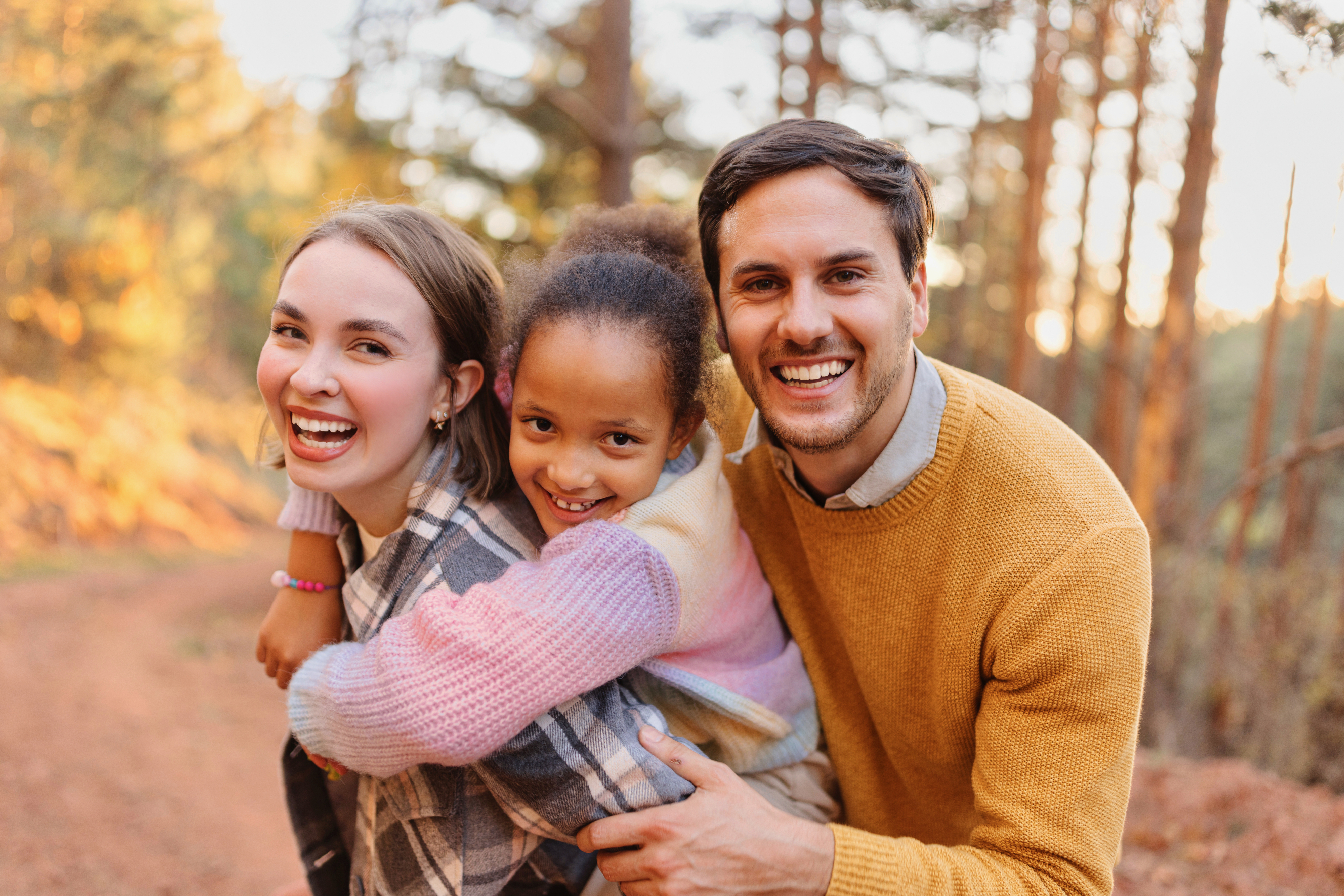 A happy family of three posing in a forest, with the child playfully on one adult&#x27;s back. All are smiling warmly at the camera