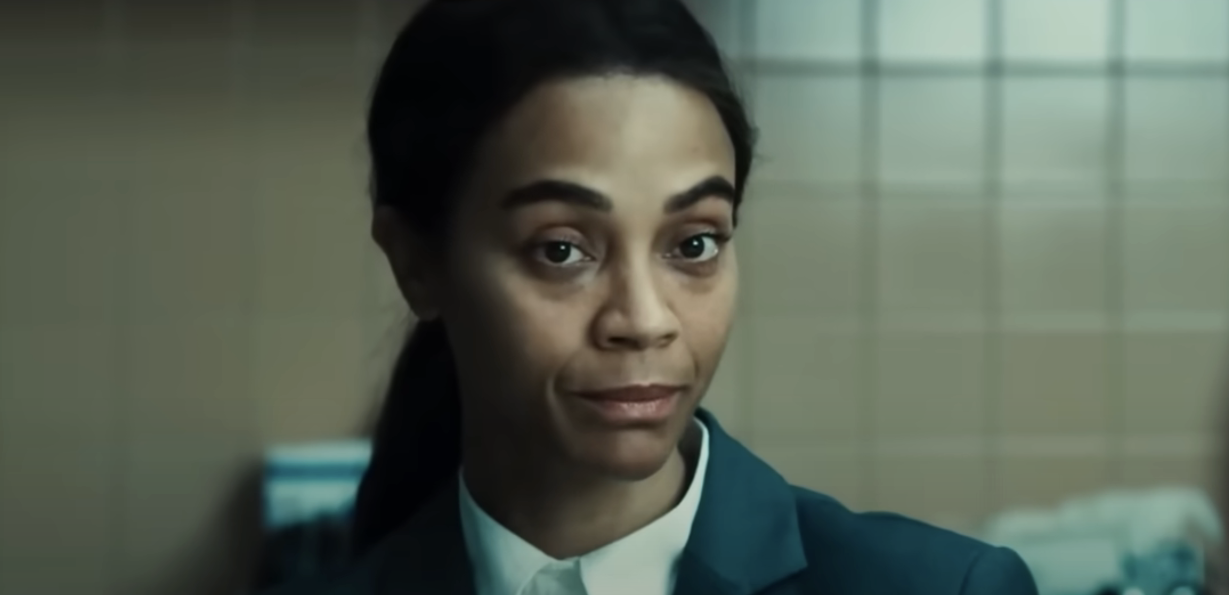 Zoe Saldana  with long dark hair tied back, wearing a collared shirt and blazer, stands in a tiled room, looking towards the camera with a neutral expression