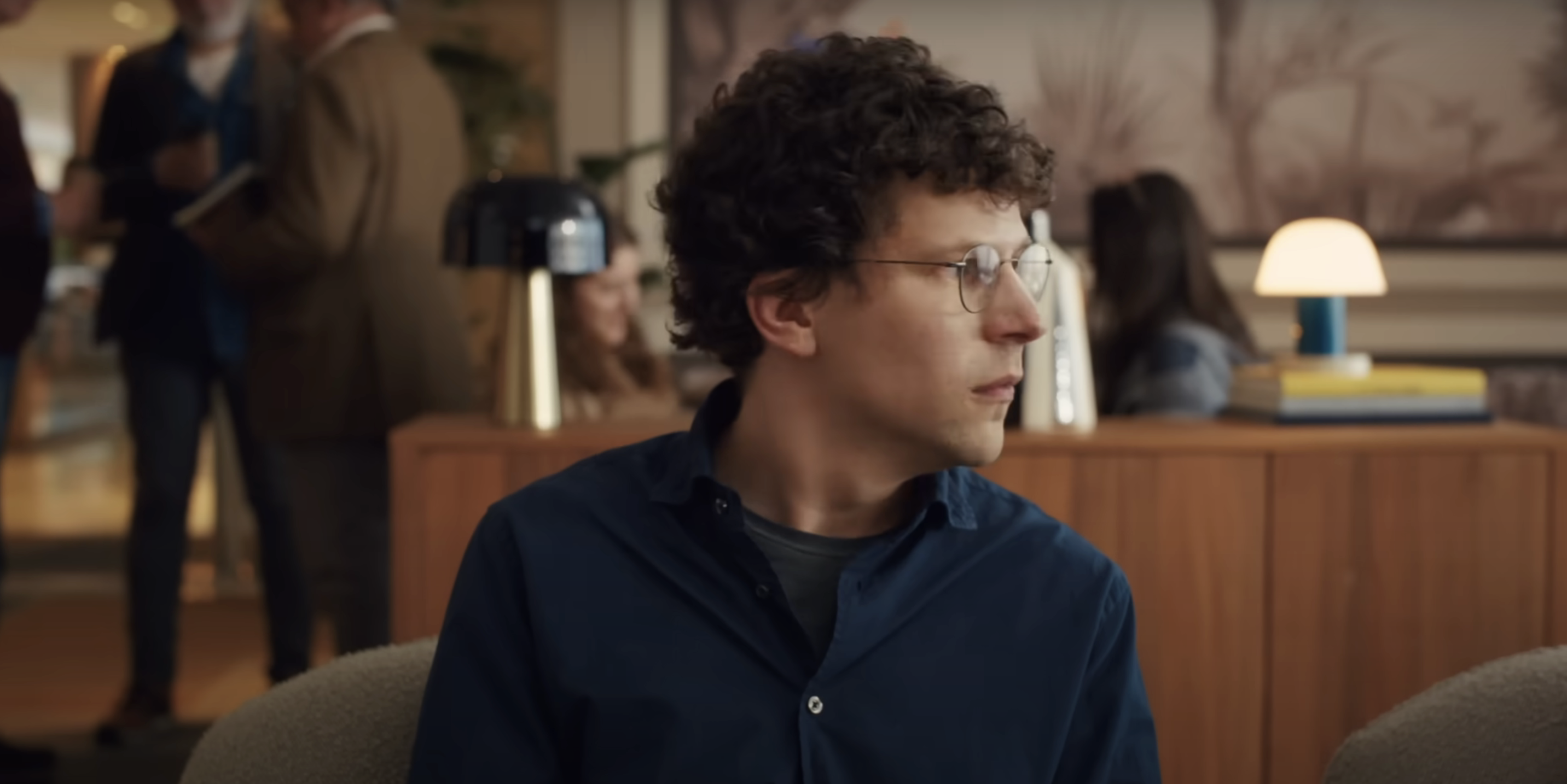 Jesse Eisenberg with glasses, curly hair, and a button-up shirt sits in an office environment, looking to the side. Other people are blurred in the background