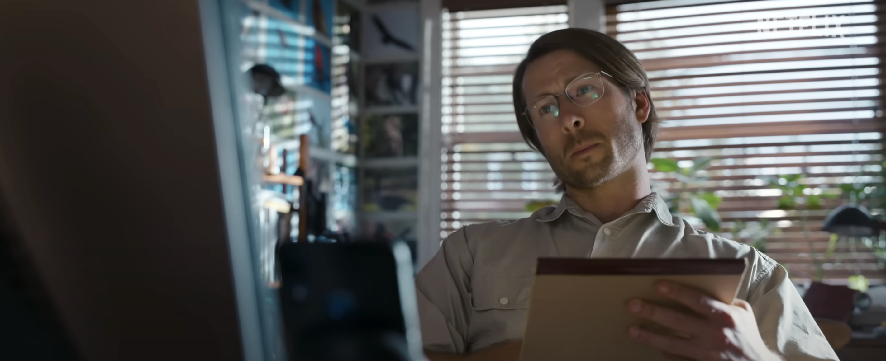 Glen Powell with glasses holding a tablet, seated in a light-filled room with plants and photos on the wall