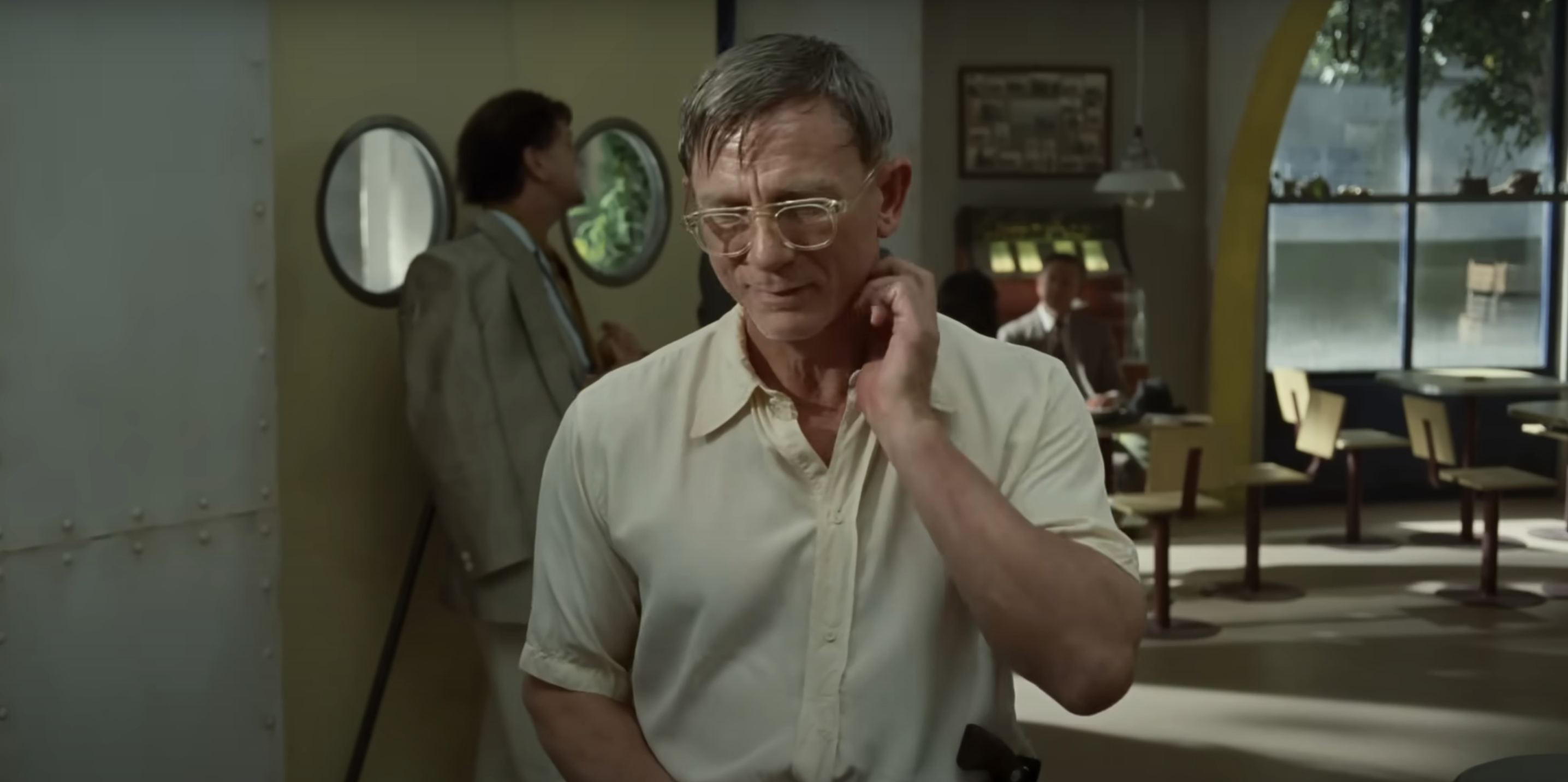 Daniel Craig in casual shirt with glasses stands in a retro-style café, touching his neck, while another person in the background looks around