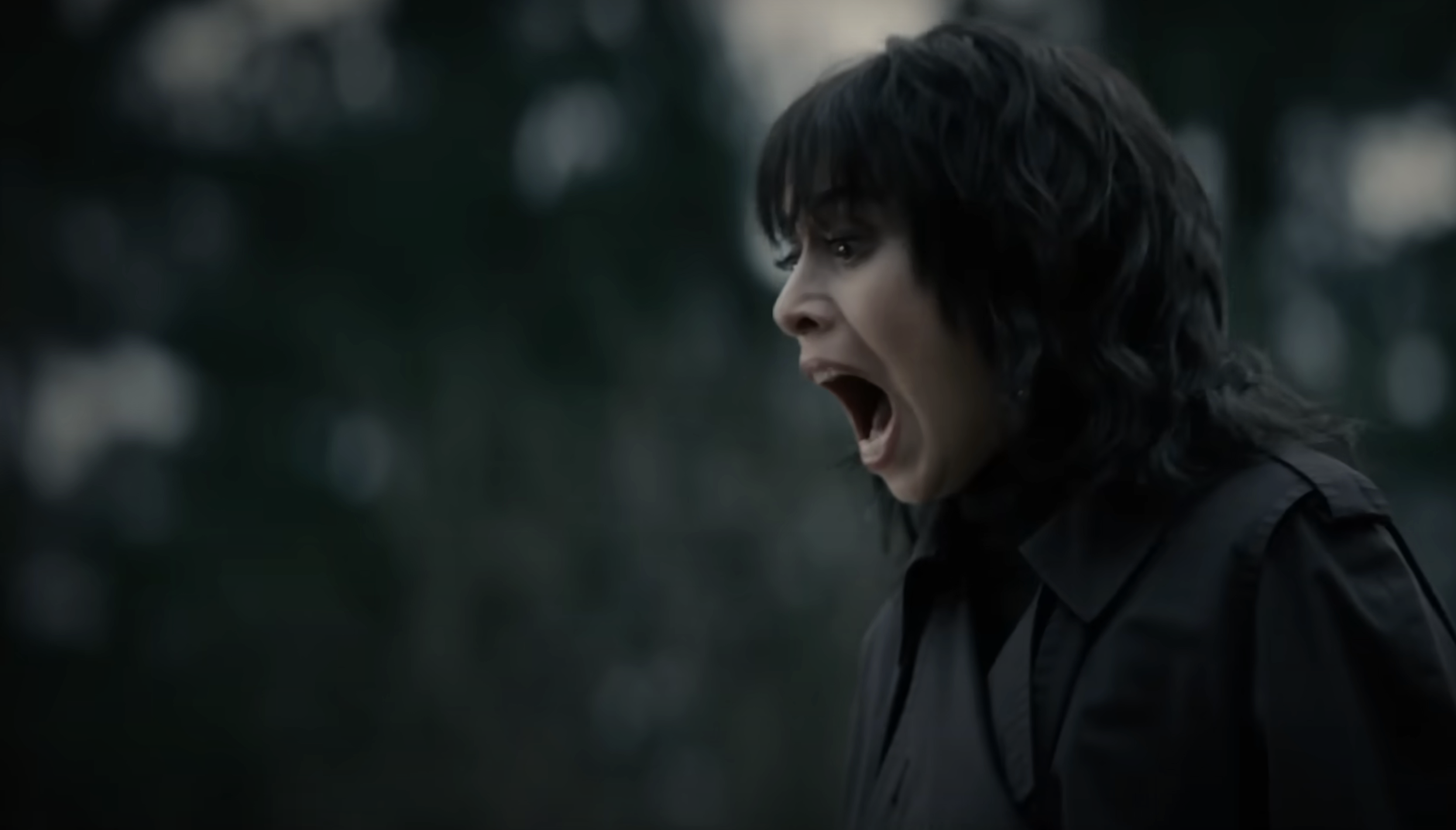 Cristin Milioti  outdoors with open mouth, wearing dark clothing, appears to be shouting or reacting emotionally. Blurred background of trees