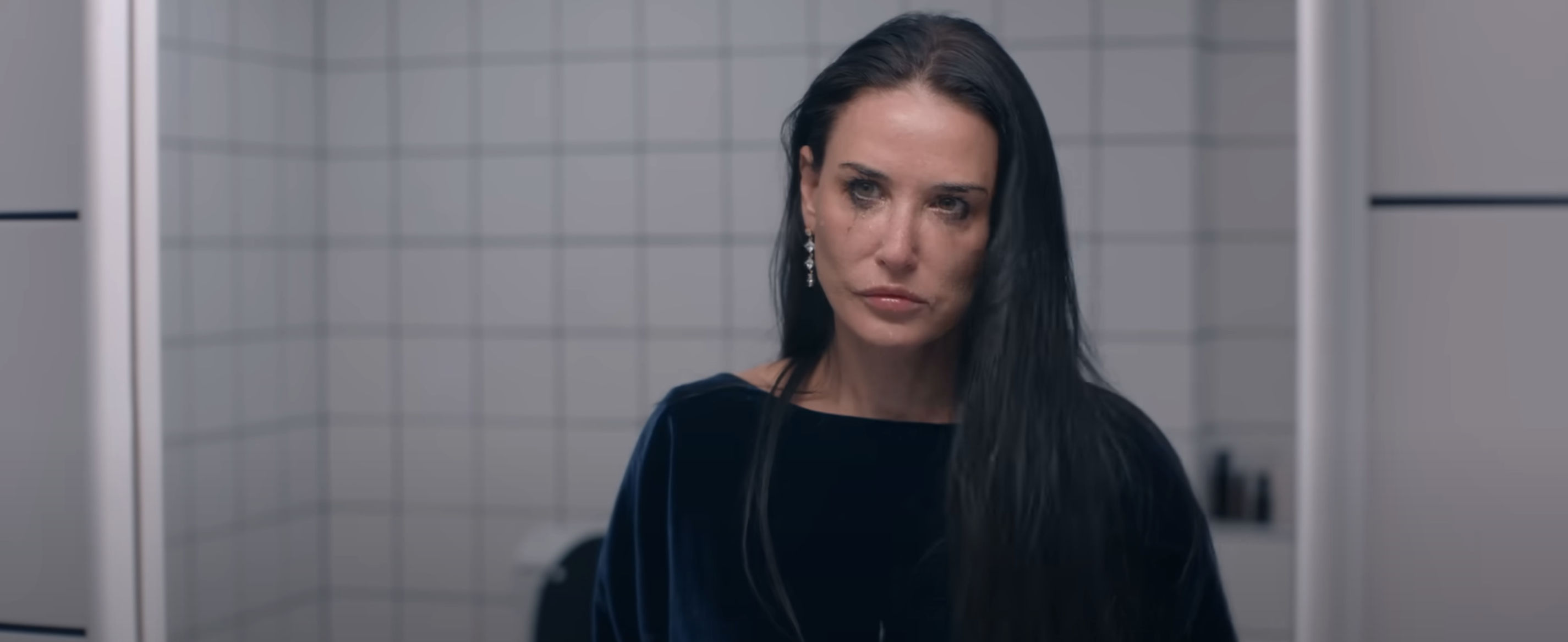 Demi Moore with long hair looks serious in a tiled bathroom. They are wearing a velvet top and earrings