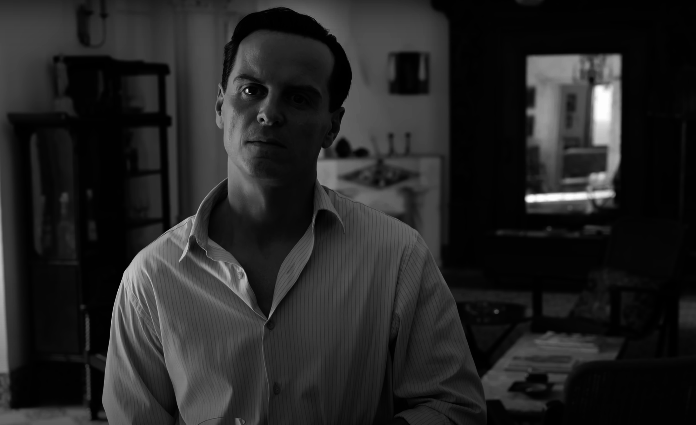 Andrew Scott stands indoors, wearing an open-collared shirt, looking thoughtfully into the distance. The room is dimly lit and has a classic style