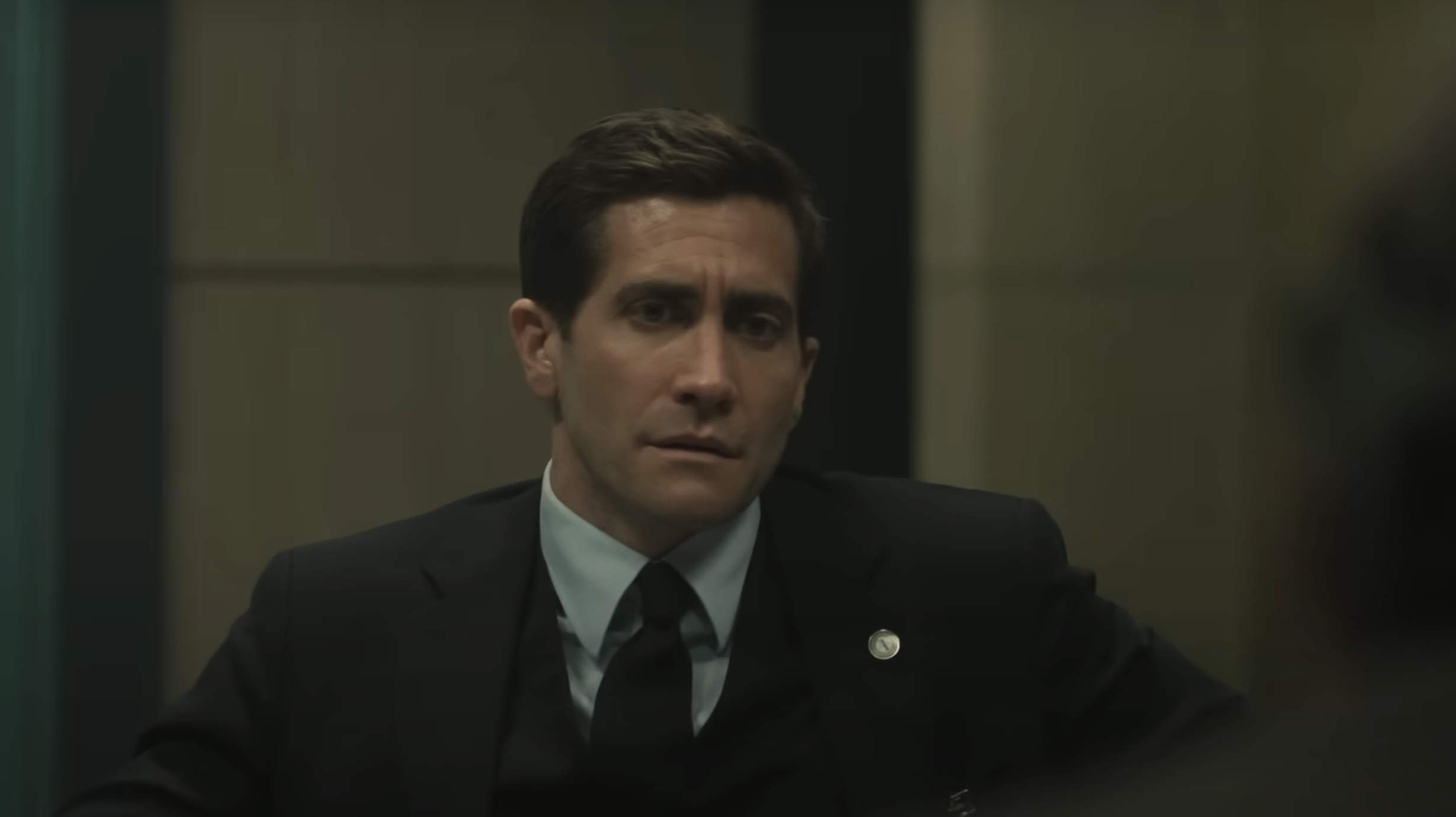 Jake Gyllenhaal in a suit and tie sits in an office, looking serious and focused