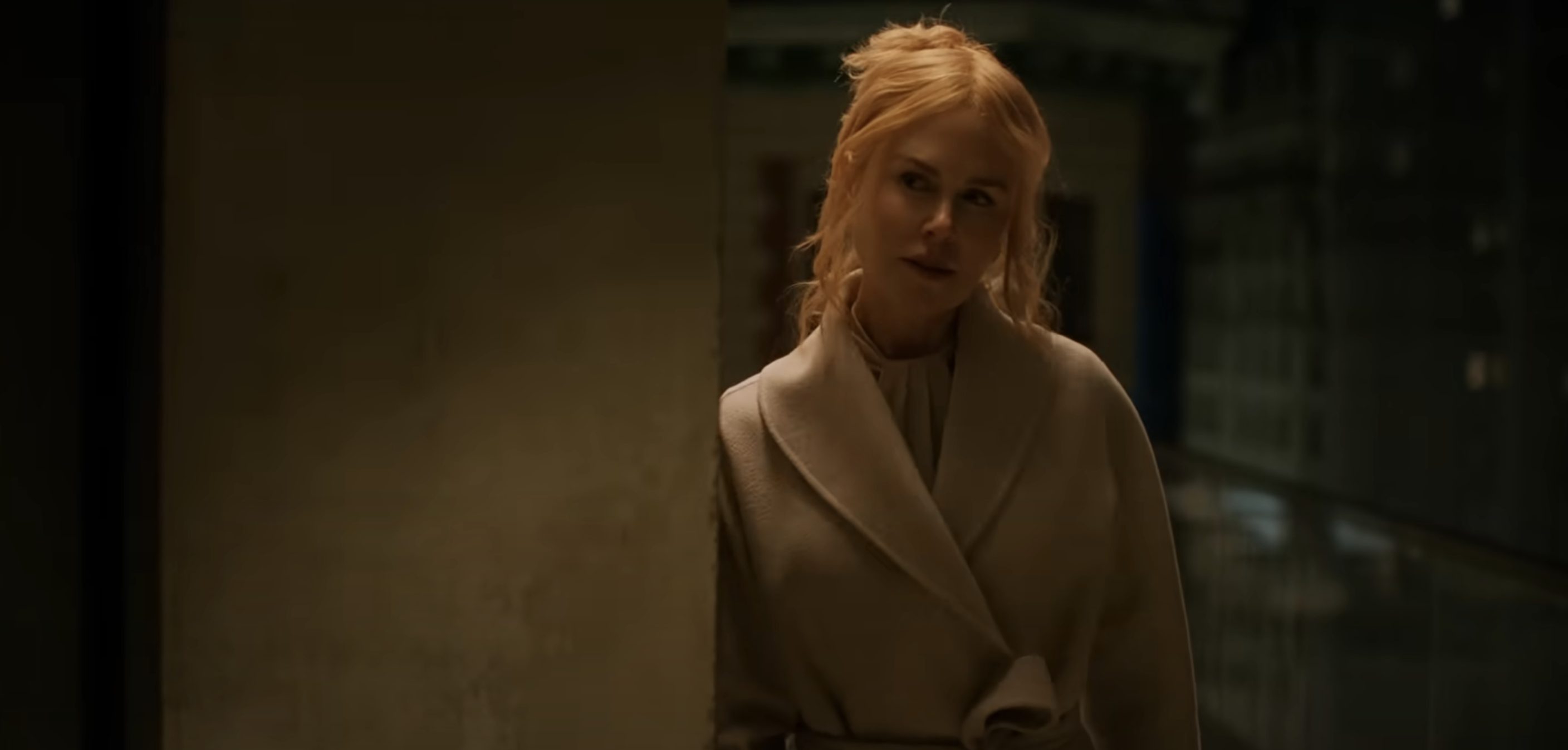 Nicole Kidman in a light coat stands by a wall, looking contemplative in a dimly lit interior setting