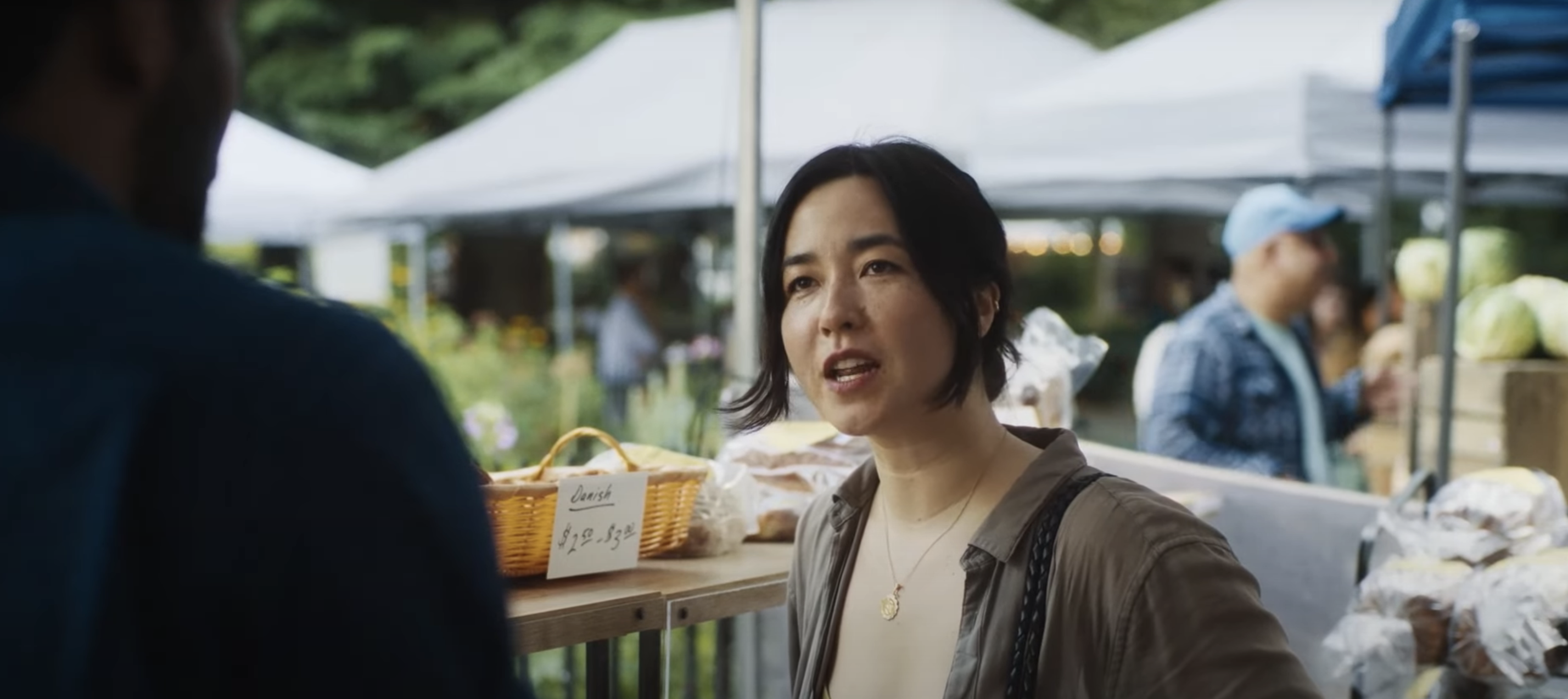 Maya Erskine converses with a man at an outdoor market, standing near a table with baked goods and price tags in the background