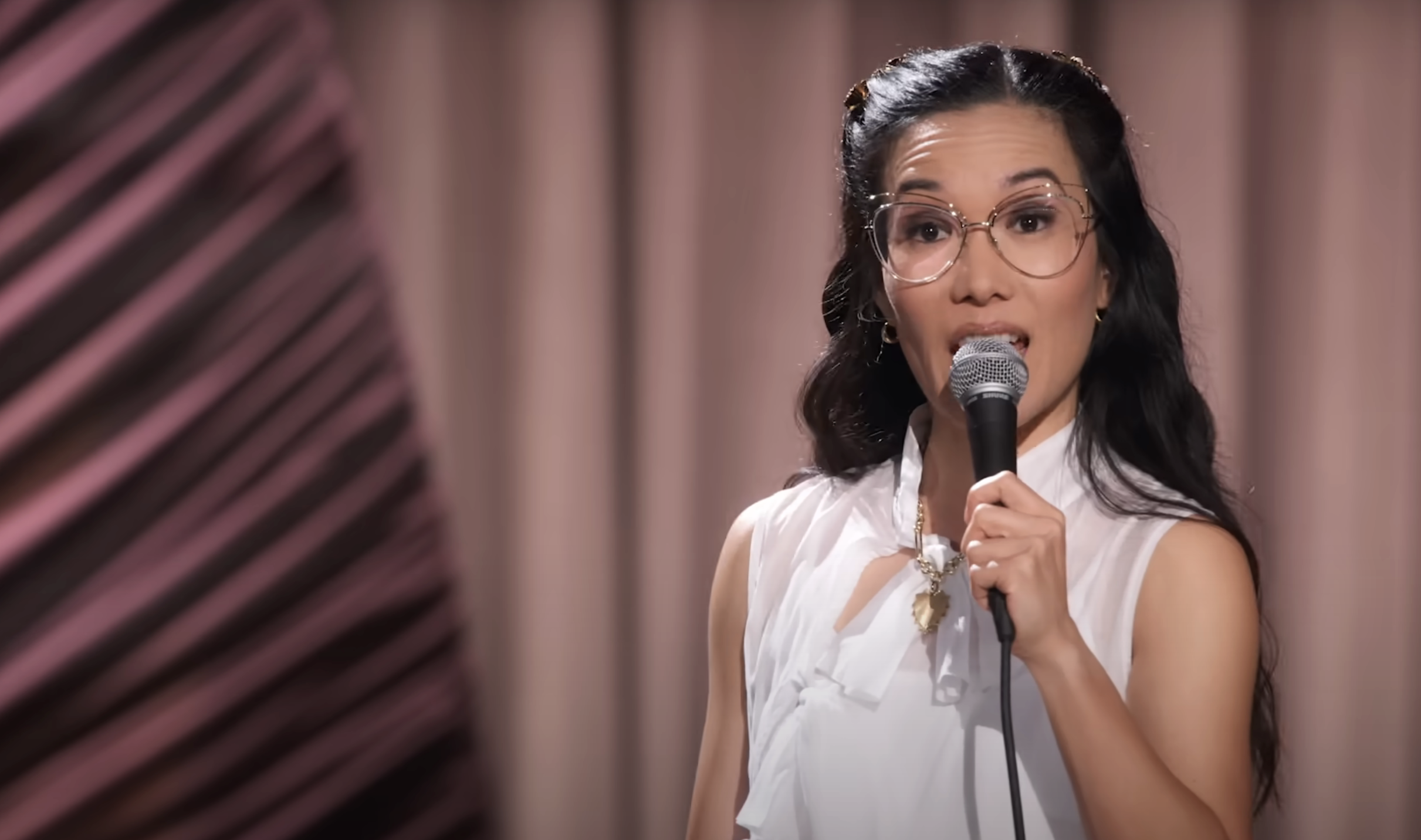 Ali Wong in white sleeveless blouse holding a microphone, speaking on stage, with a background featuring vertical lines