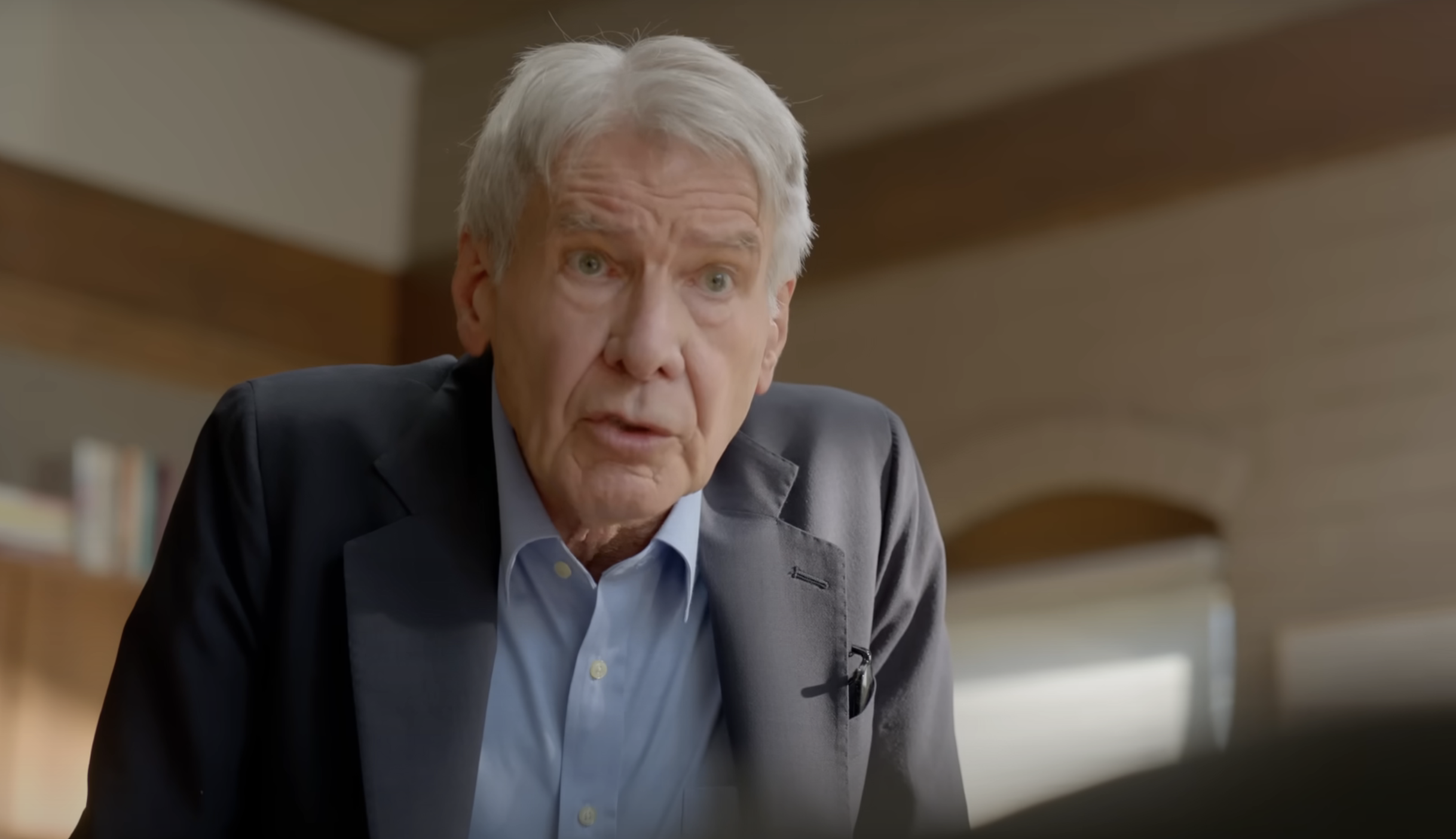 Harrison Ford in a suit, leaning forward, in an interior setting with books in the background