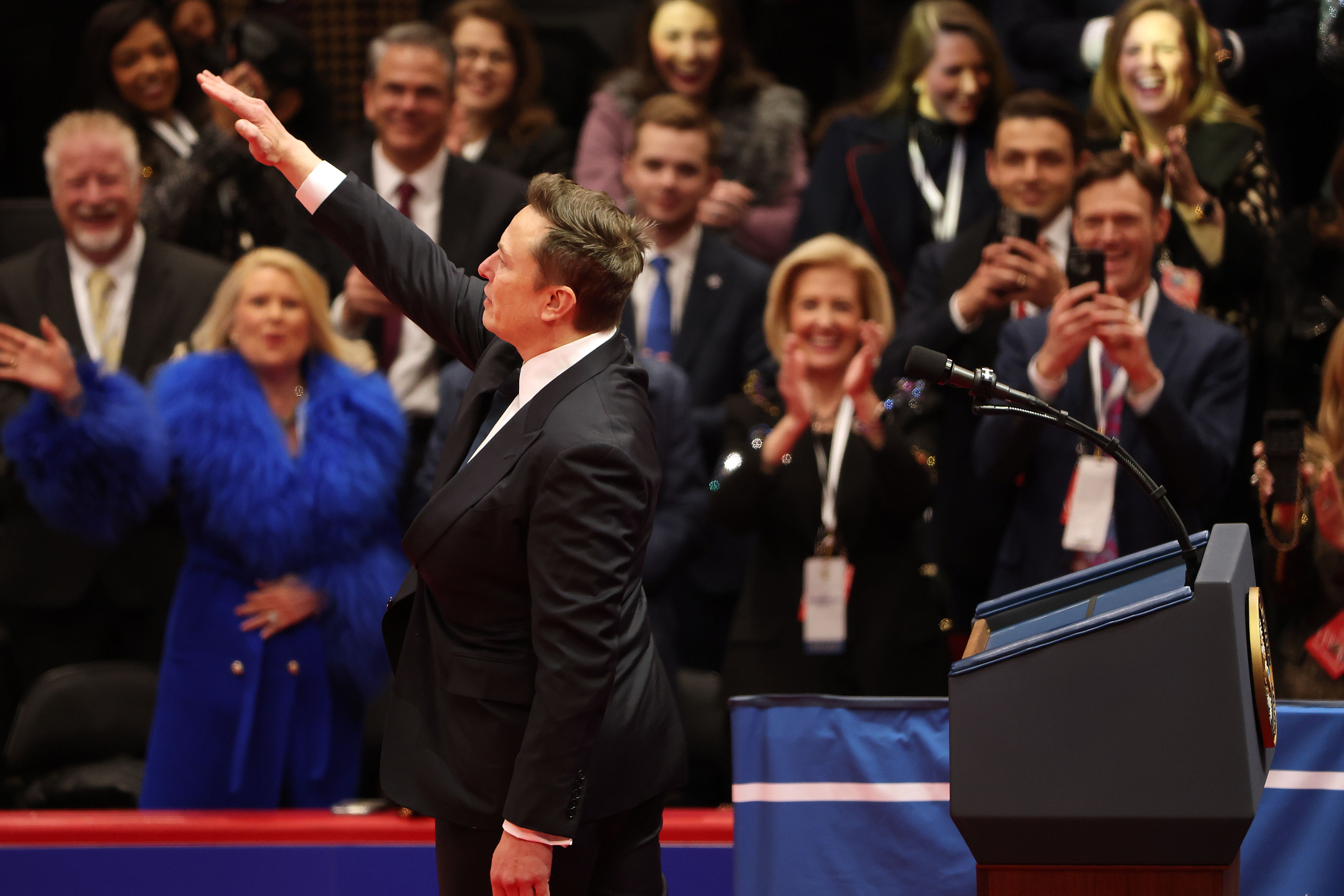 A person in a suit gestures to a cheering audience from a podium at an event. There&#x27;s a lively crowd celebrating in the background