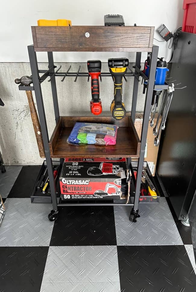 A rolling utility cart with power tools, a towel set, and contractor trash bags neatly organized on it. The cart is placed in a garage or workshop environment