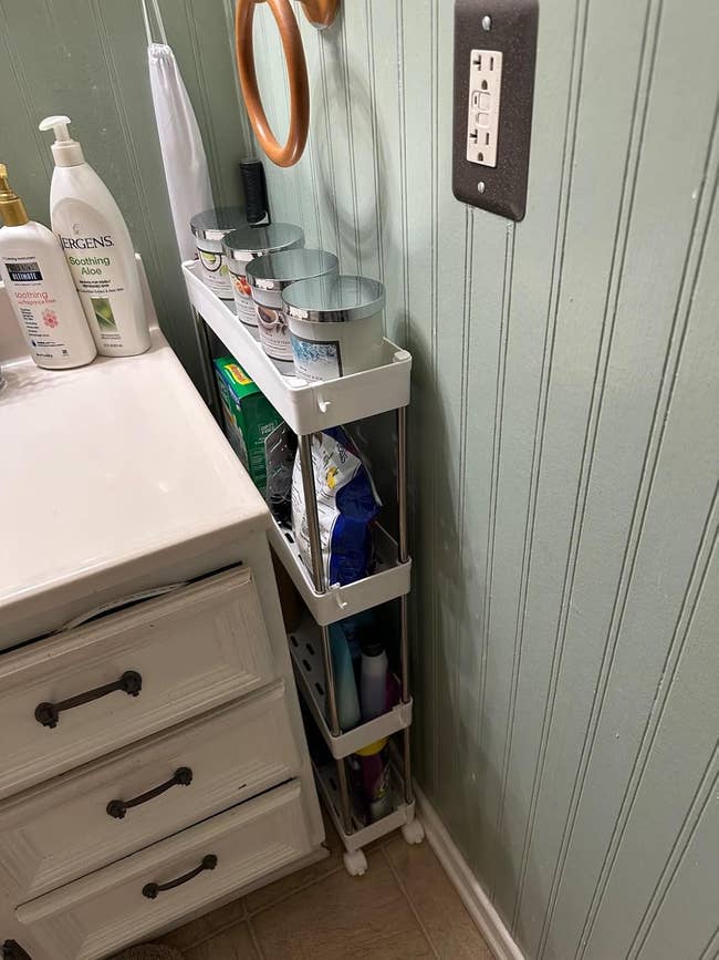 Bathroom storage rack next to a dresser, holding toiletries like bottles, cans, toiletries, and cleaning products