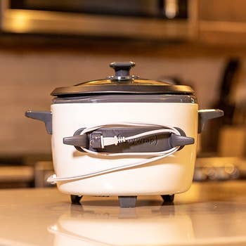 A gray oval-shaped rubber adhesive with two prongs attached to a slow cooker with the cord wrapped around it 