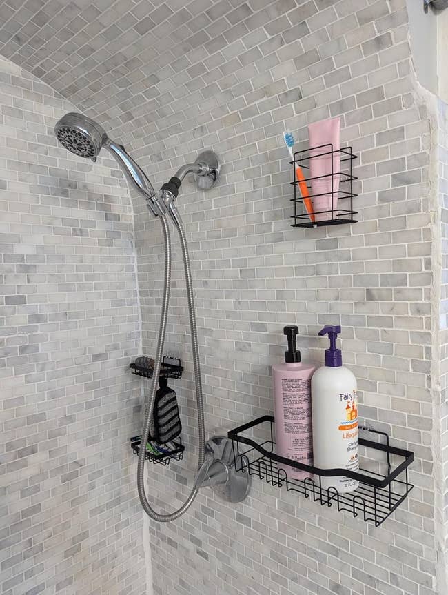 A shower with a handheld showerhead, a wire shelf holding body wash and shampoo, and a wall-mounted toothbrush holder with a toothbrush and toothpaste