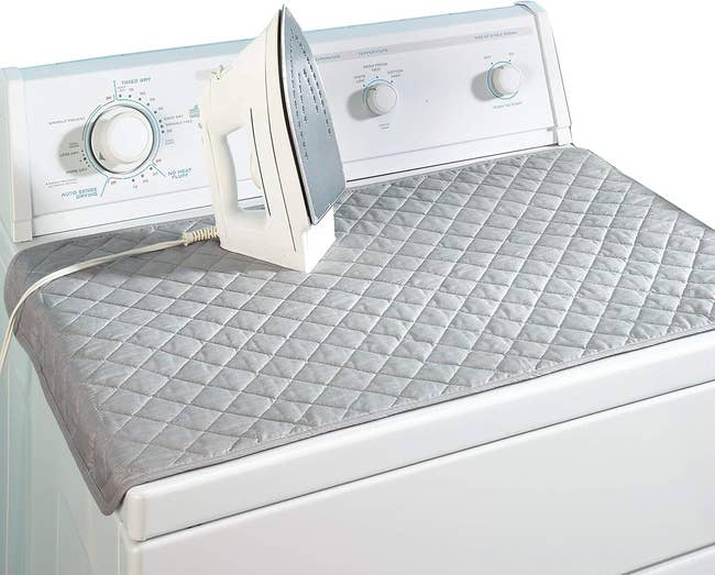 Washer or dryer with quilted gray ironing pad on top, white iron placed on the pad. Controls and settings of the appliance are visible in the background