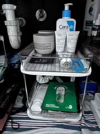 Skincare products arranged on a two-tier shelf under a sink, including CeraVe lotion, Olaplex No.3 and No.8, and COSRX Cica Calm Mask