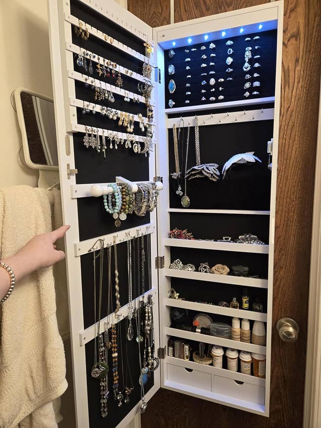 A wall-mounted jewelry organizer is shown open, displaying various rings, necklaces, bracelets, and other accessories. Shelves at the bottom hold beauty products