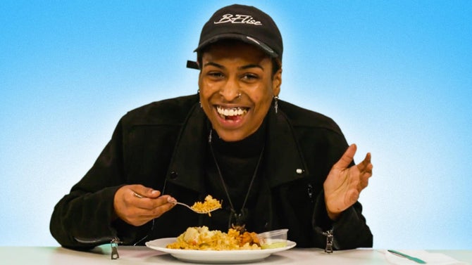 A contestant in a black cap smiles and shrugs holding a forkful of rice with a plate of soul food in front of them.