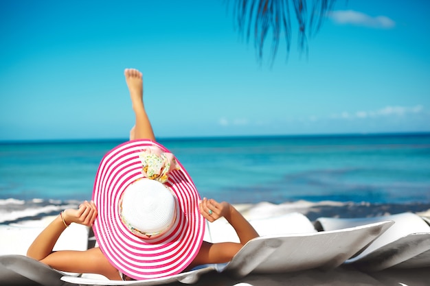 Foto gratuita modello di bella donna che prende il sole sulla sedia di spiaggia in bikini bianco in cappellino da sole variopinto dietro l'oceano blu dell'acqua di estate