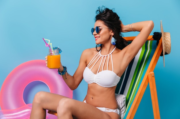 Foto gratuita donna carina che beve cocktail e gioca con i capelli studio shot di allegra ragazza abbronzata in bikini seduta su chaise longue