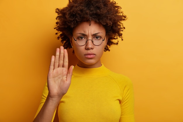 Foto gratuita la giovane donna dai capelli ricci scontenta tiene il palmo della mano, fa il gesto di proibire, limita qualcosa, guarda seriamente, indossa gli occhiali, maglione giallo, posa al coperto, ha un'espressione di avvertimento