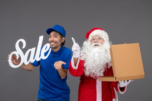 Foto gratuita vista frontale di babbo natale con corriere maschio che tiene banner di vendita e scatola di cibo sul muro grigio