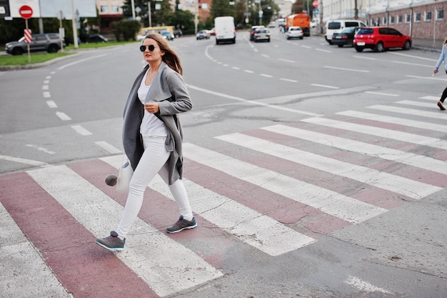 Foto gratuita ragazza in cappotto grigio con occhiali da sole e borsetta che cammina sul passaggio pedonale