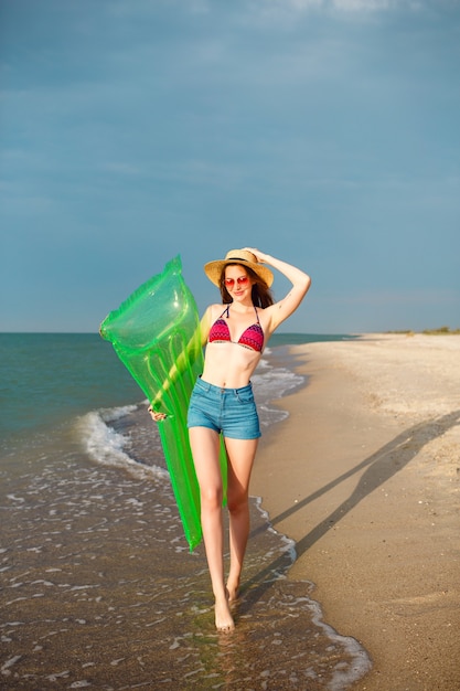 Foto gratuita donna graziosa felice che si diverte sulla spiaggia, indossa abbigliamento da spiaggia elegante, cappello bikini e pantaloncini di jeans, gambe lunghe, corpo aderente, tenendo il materasso ad aria e camminando vicino all'oceano