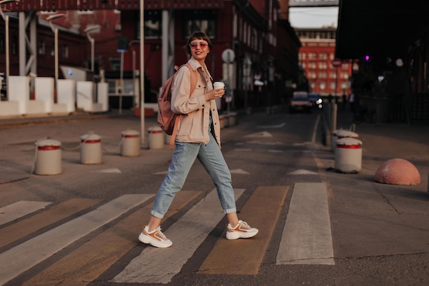 Foto gratuita la signora ottimista in abito leggero cammina fuori ragazza moderna in jeans sneakers occhiali e giacca beige con zaino tiene una tazza di tè in strada