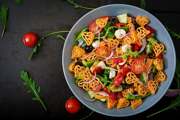 Foto gratuita insalata di pasta a forma di cuore con pomodori, cetrioli, olive, mozzarella e cipolla rossa alla greca. disteso. vista dall'alto